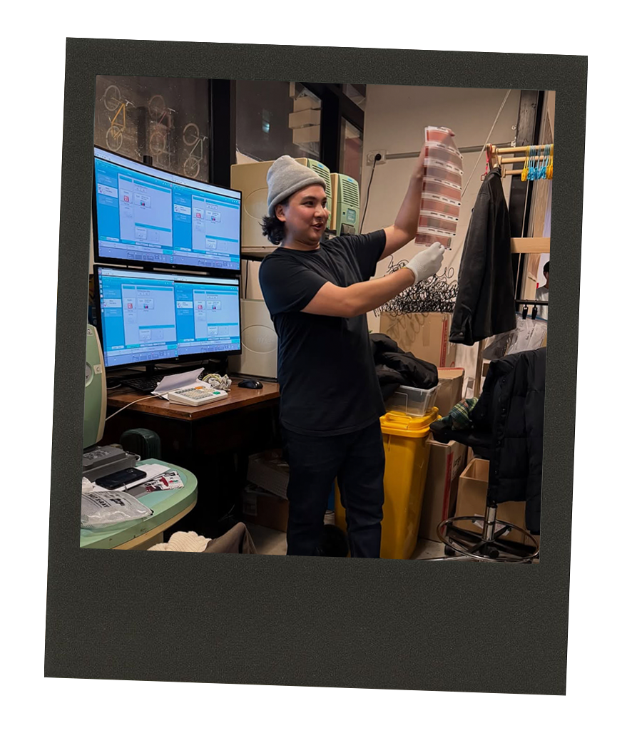 A person wearing a gray beanie and black t-shirt holds a swatch of fabric or material in a cluttered workspace with multiple computer monitors, office supplies, and miscellaneous items.