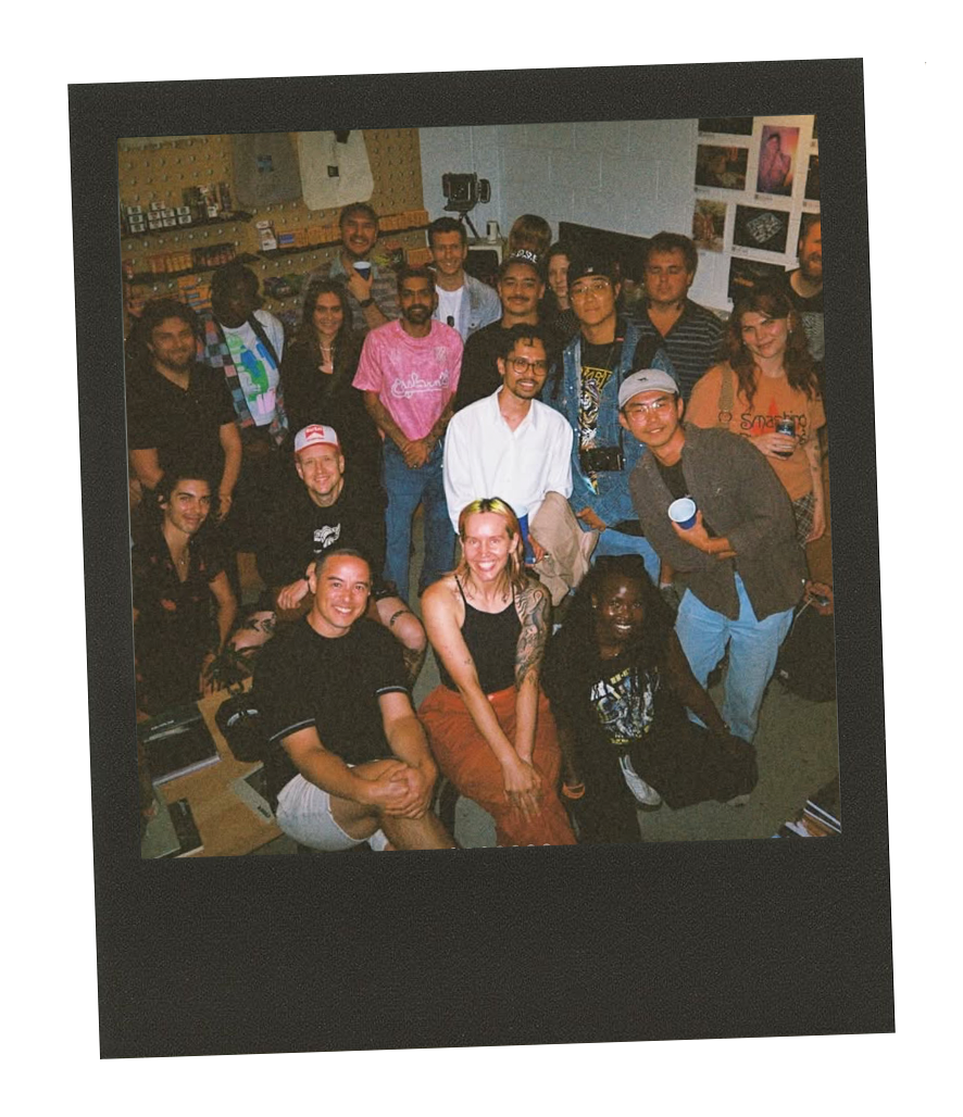 Group of diverse people gathered indoors for a photo, some smiling, with artwork on the walls and a camera on a tripod in the background.