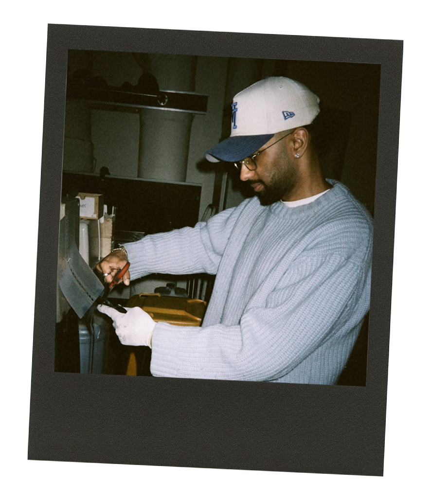 A man wearing a white cap, glasses, and a gray sweater is working with tools at a workspace with a computer and office supplies.
