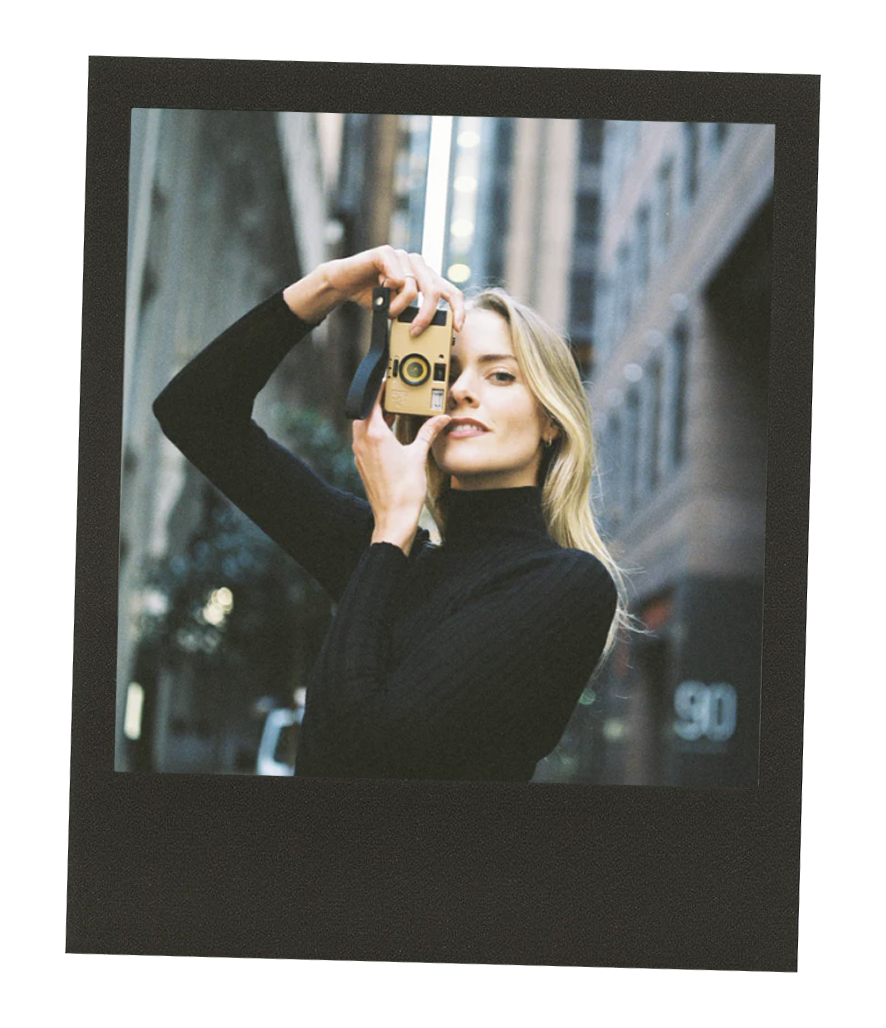 A woman with blonde hair wearing a black turtleneck taking a photo with a yellow camera on a city street.