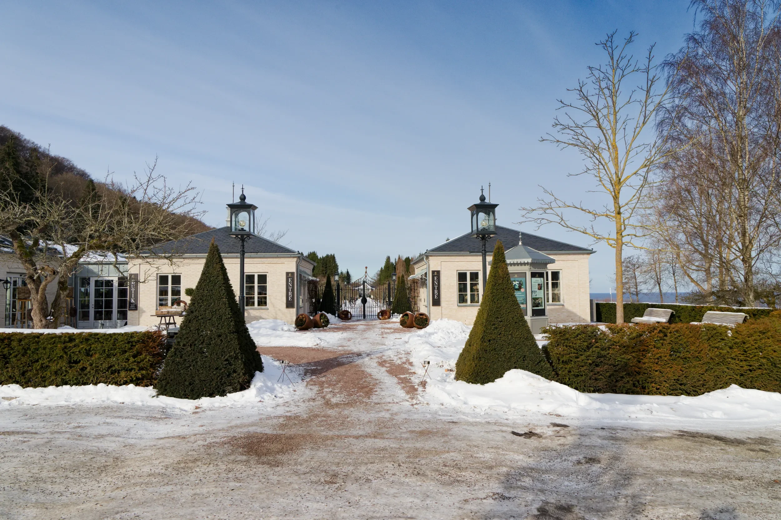 Ingången till Norrvikens trädgårdar i Båstad, omgivna av träd och buskar, inklusive två ljusstakar med lampor längs vägen.