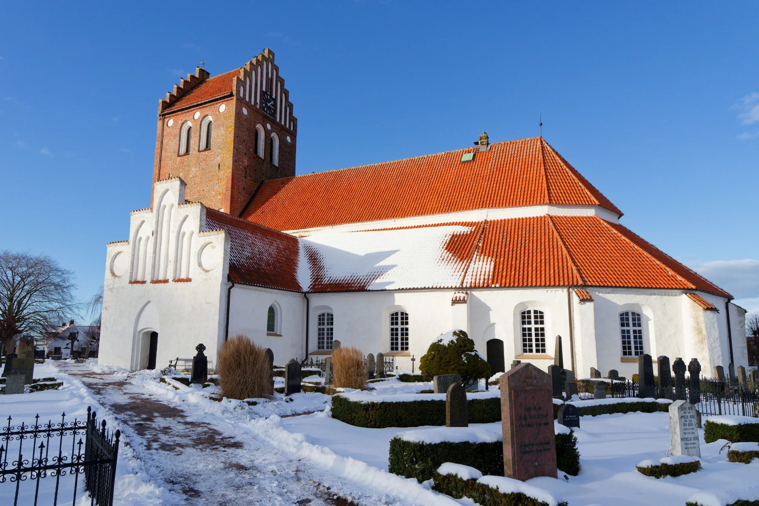 Mariakyrkan i Båstad med röd takpanna och ett långt vitt väggparti, fakturerad i en fristående kyrktornvägg, med gravar framför och en klar blå himmel.