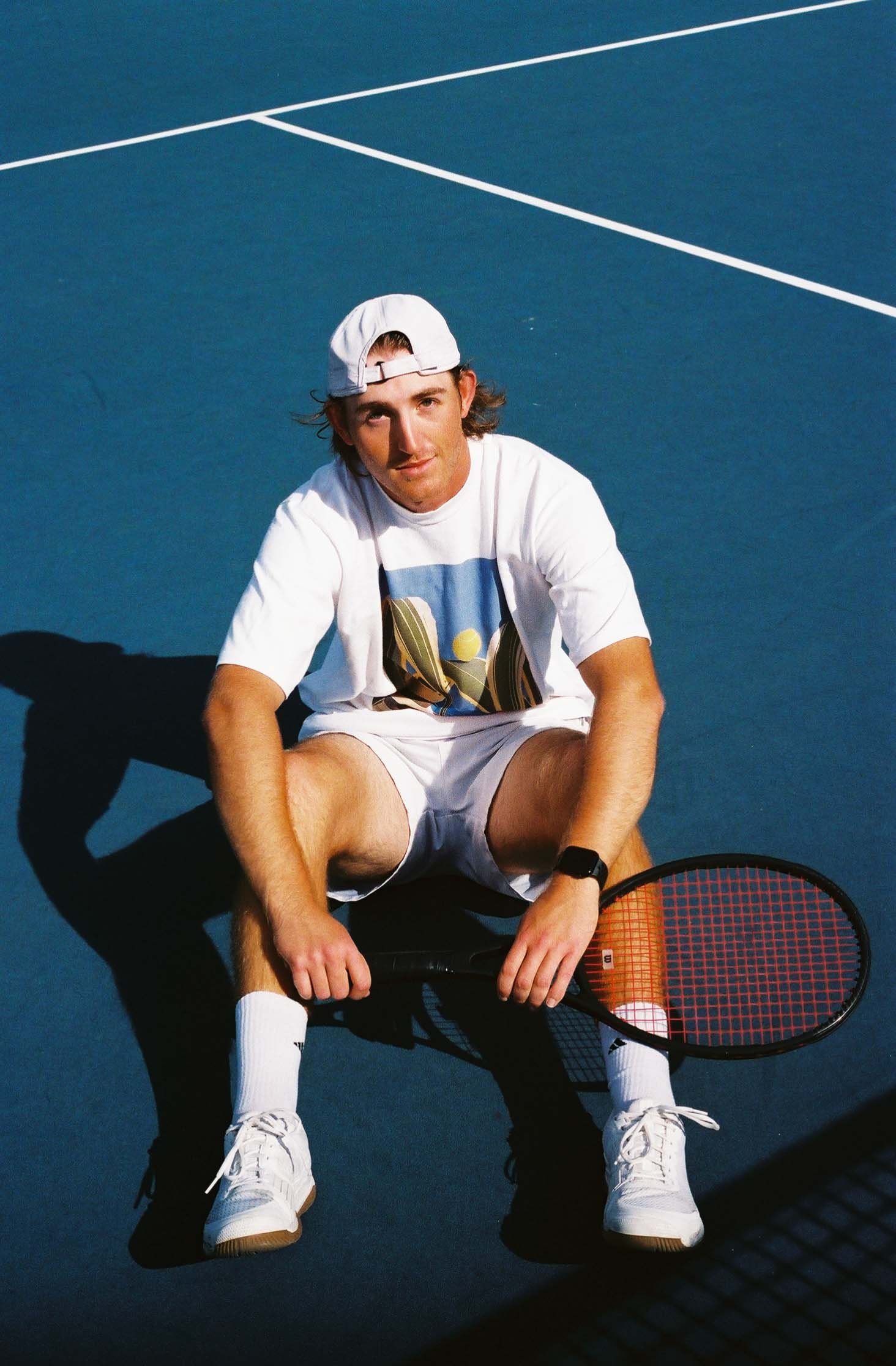 A young man with brown hair, wearing a white cap, white T-shirt with a graphic, white shorts, and white sneakers, sitting on a tennis court with a tennis racket, looking up at the camera.