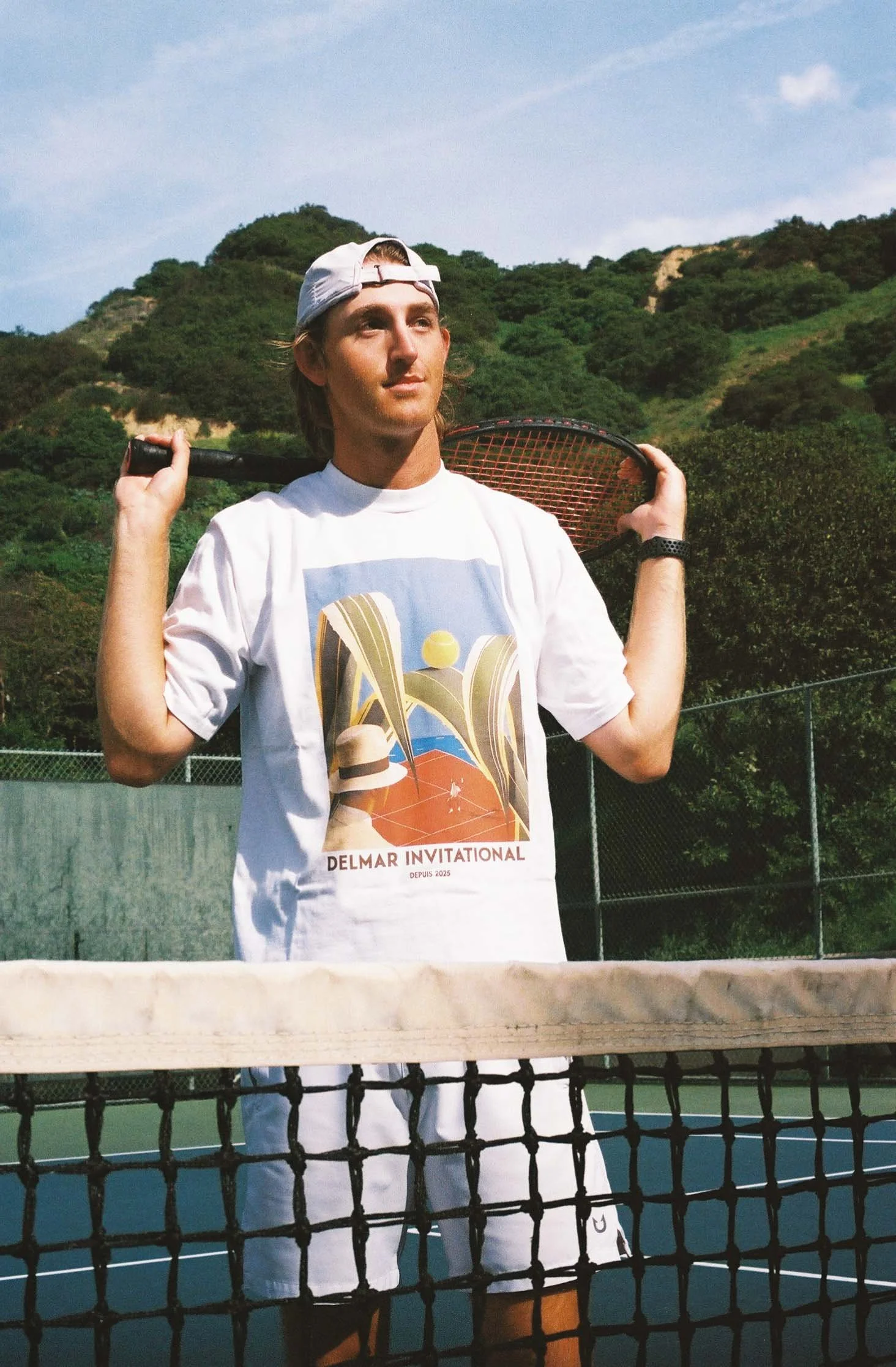 A young man standing on a tennis court, holding a tennis racket over his shoulders, wearing a white cap backward and a graphic T-shirt that says "Delmar Invitational." He is outdoors with green hills and a partly cloudy sky in the background.