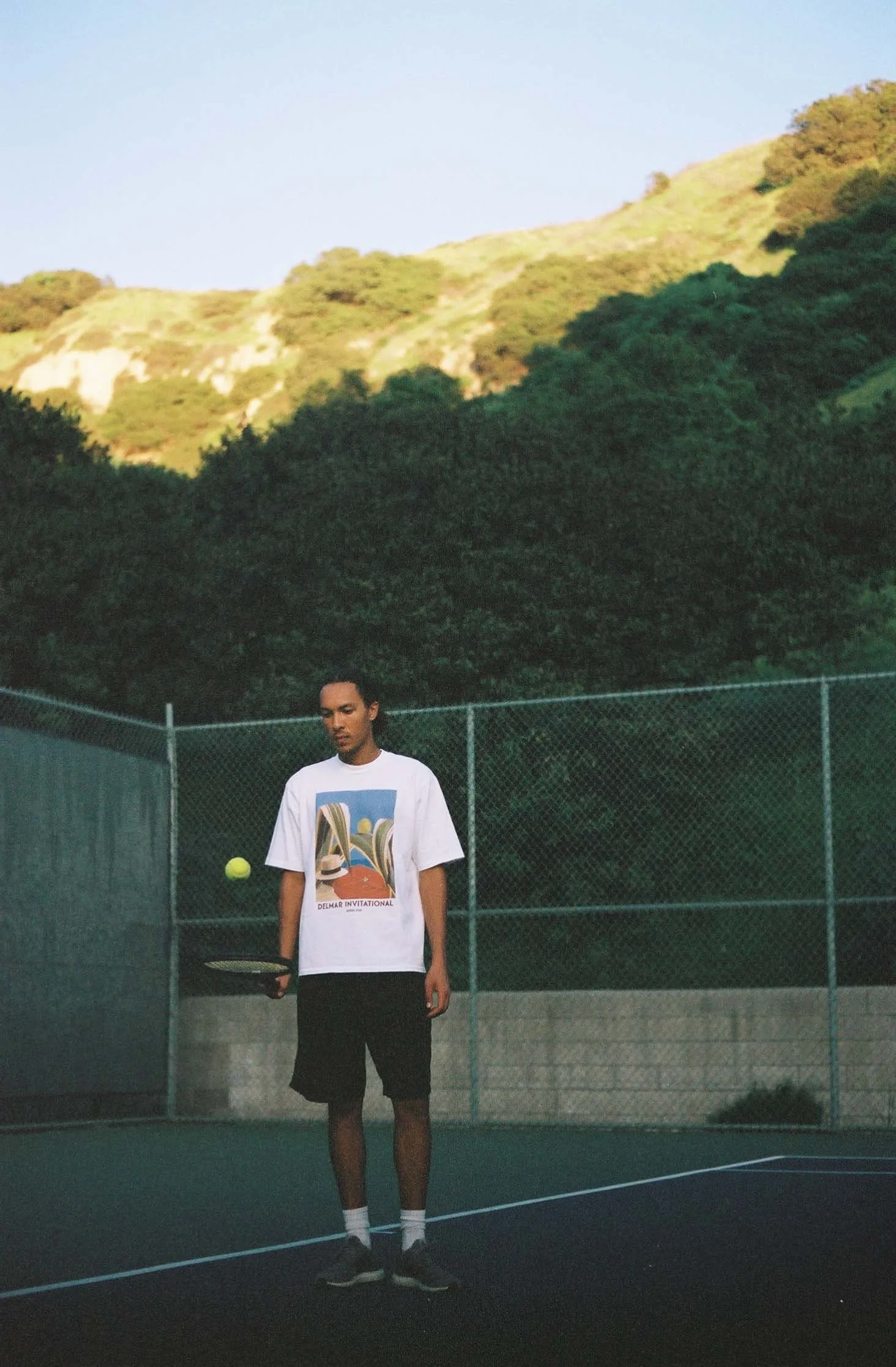 A young man on a tennis court holding a racket, with a tennis ball mid-air. He is wearing a white graphic t-shirt, black shorts, white socks, and black shoes. The court is surrounded by a chain-link fence with a backdrop of green hills and trees.