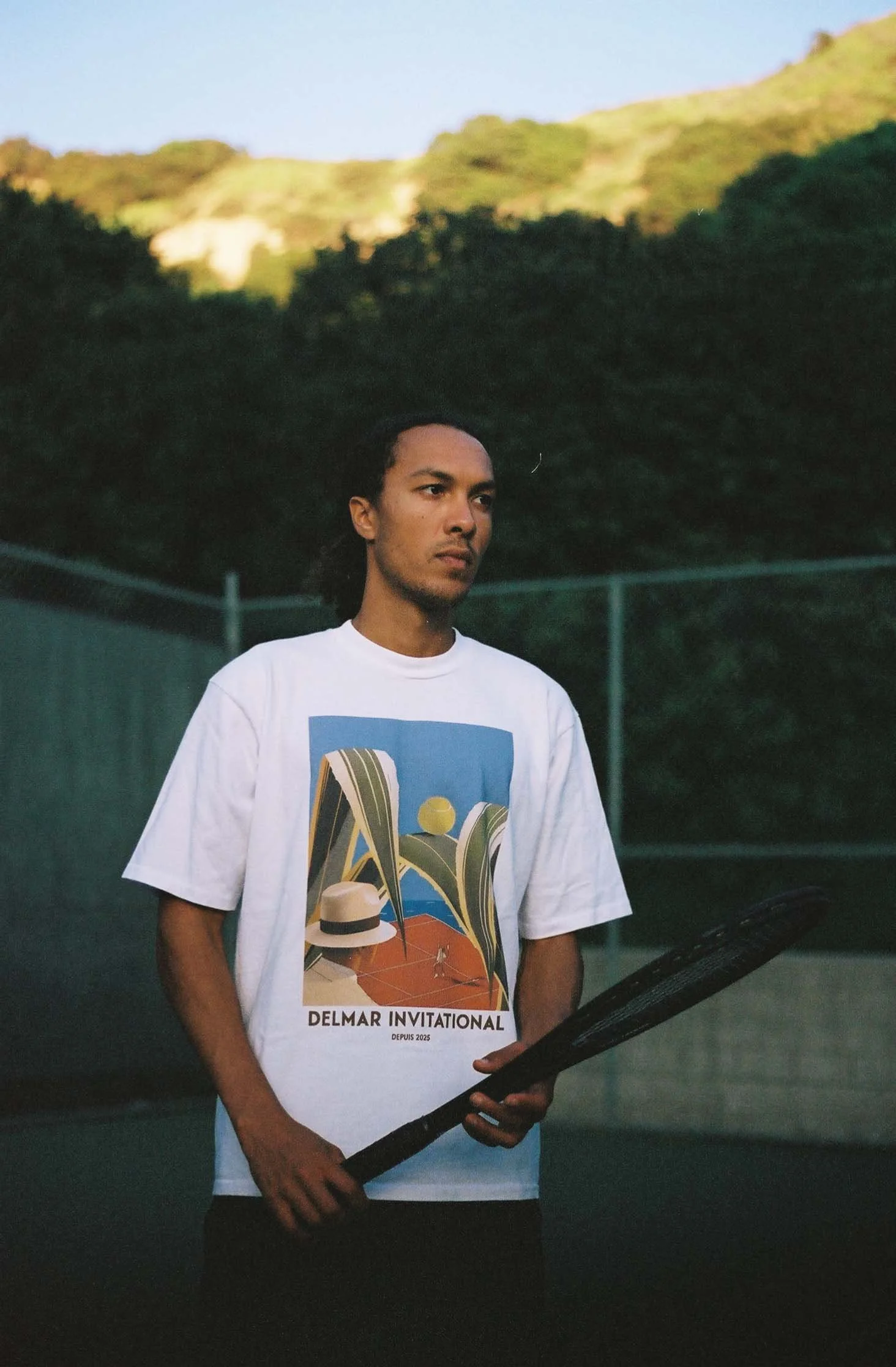 A man standing on a tennis court at dusk, holding a tennis racket, wearing a white T-shirt with colorful graphic design and text that reads "Delmar Invitational," with dark trees and hills in the background.
