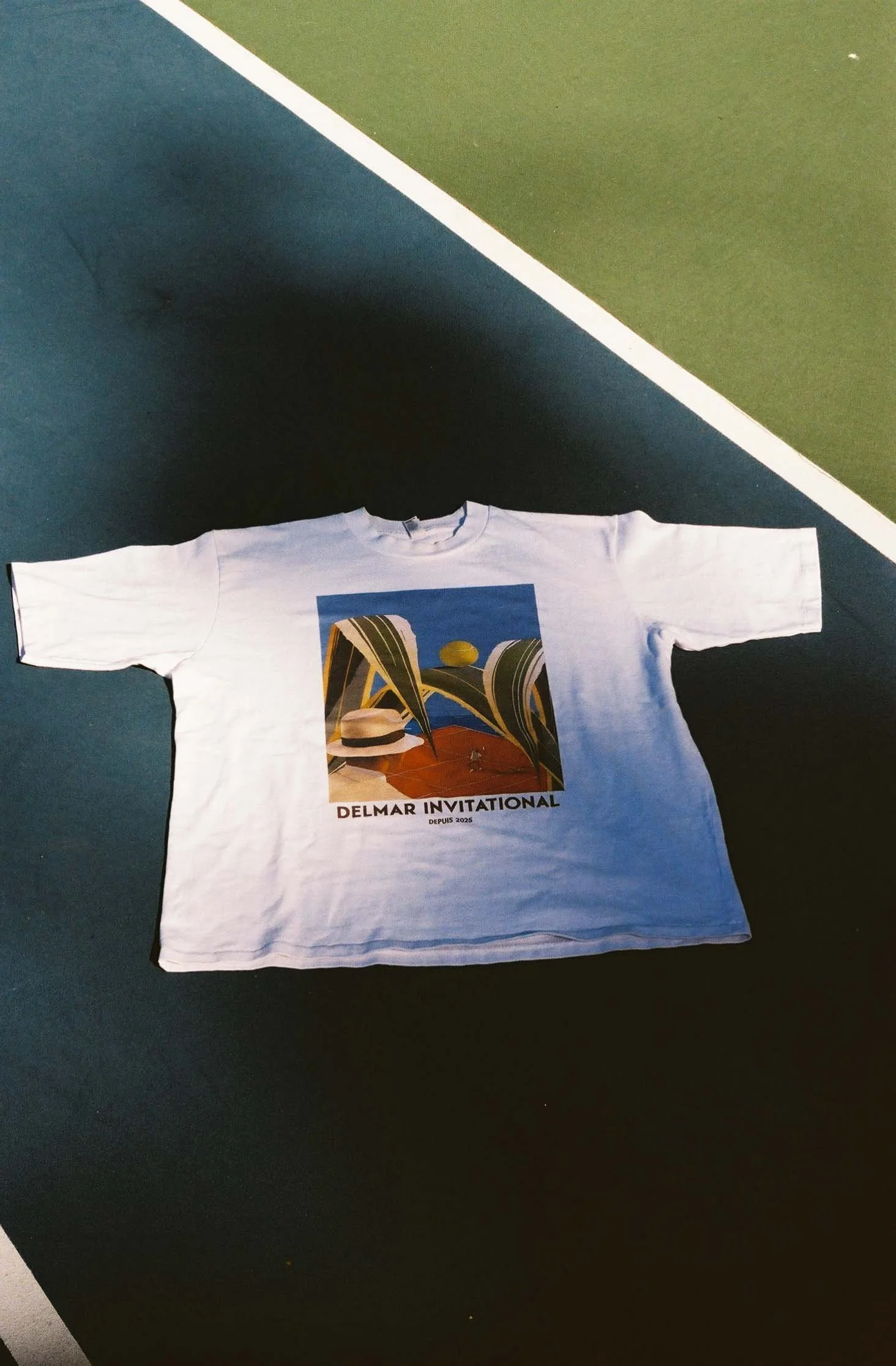 A white T-shirt with a graphic print of futuristic abstract architecture, a straw hat, and green and yellow orbs, with the text "Delmar Invitational" and "Depuis 2025" beneath the image, laid flat on a colorful surface.