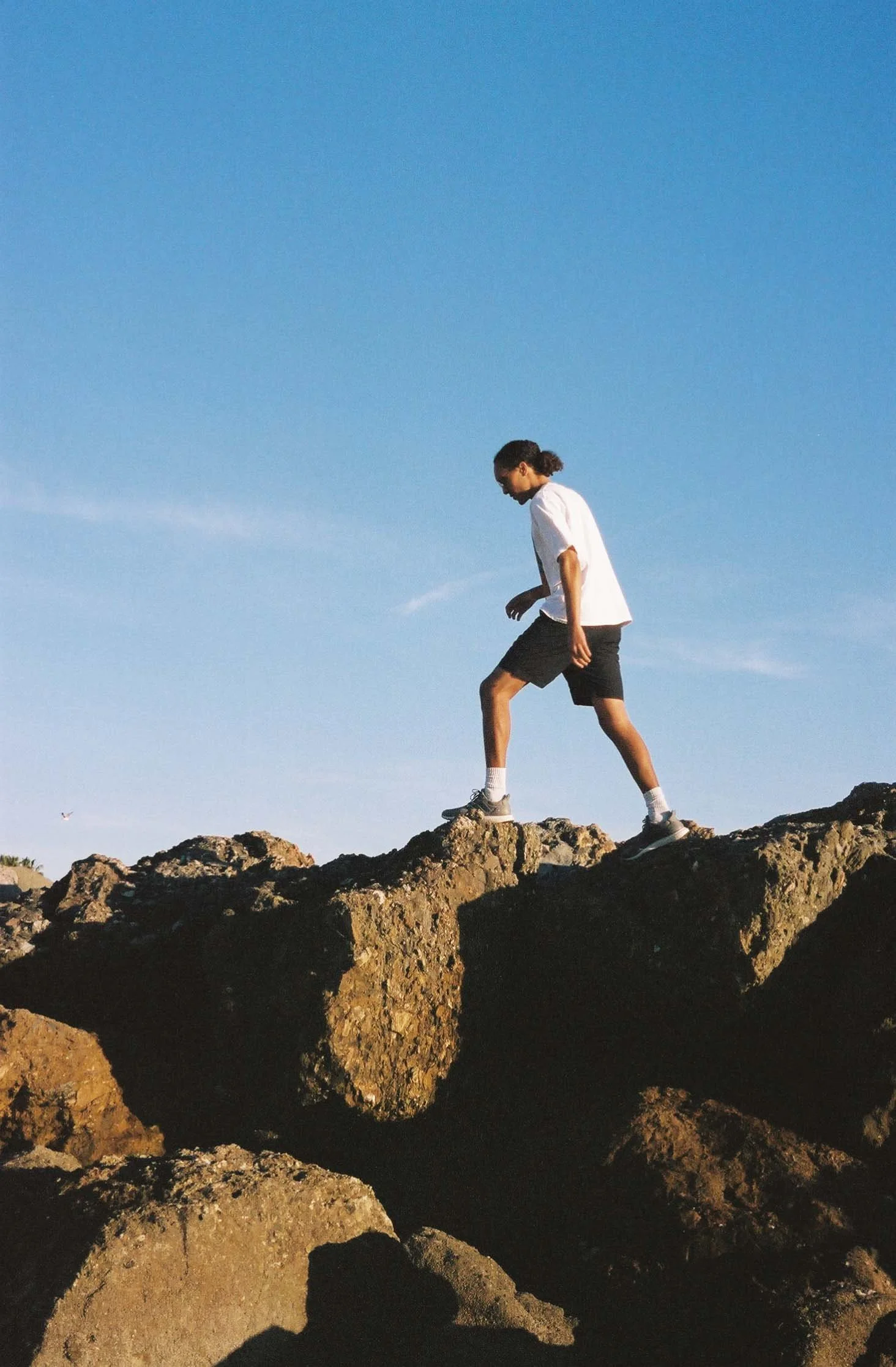 A person with long hair tied back, wearing a white shirt, black shorts, earrings, white socks, and sneakers, walking on large rocks outdoors with a clear blue sky in the background.