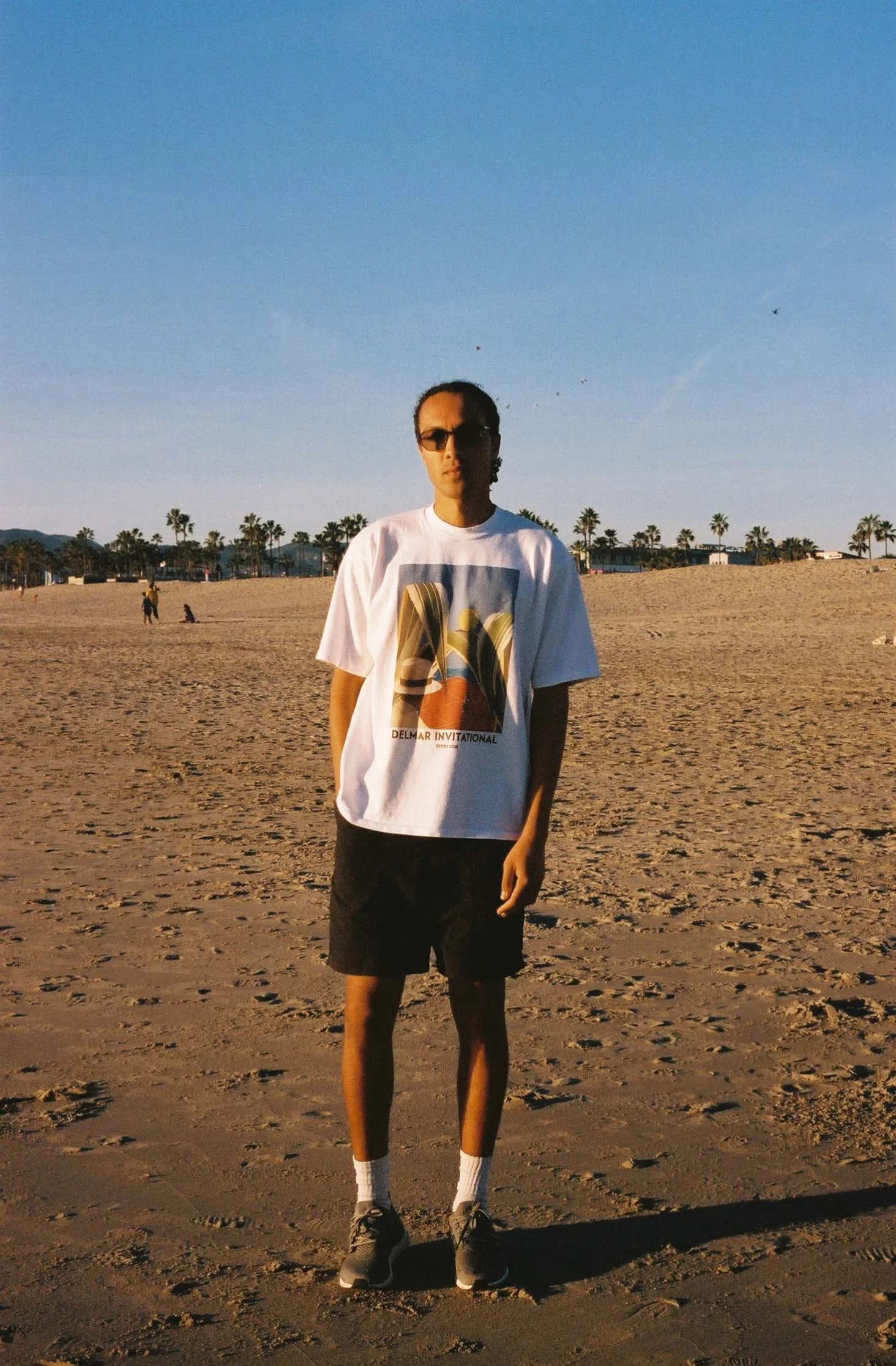 A person standing on a sandy beach with palm trees and a clear blue sky in the background, wearing sunglasses, a white graphic t-shirt, black shorts, white socks, and sneakers.