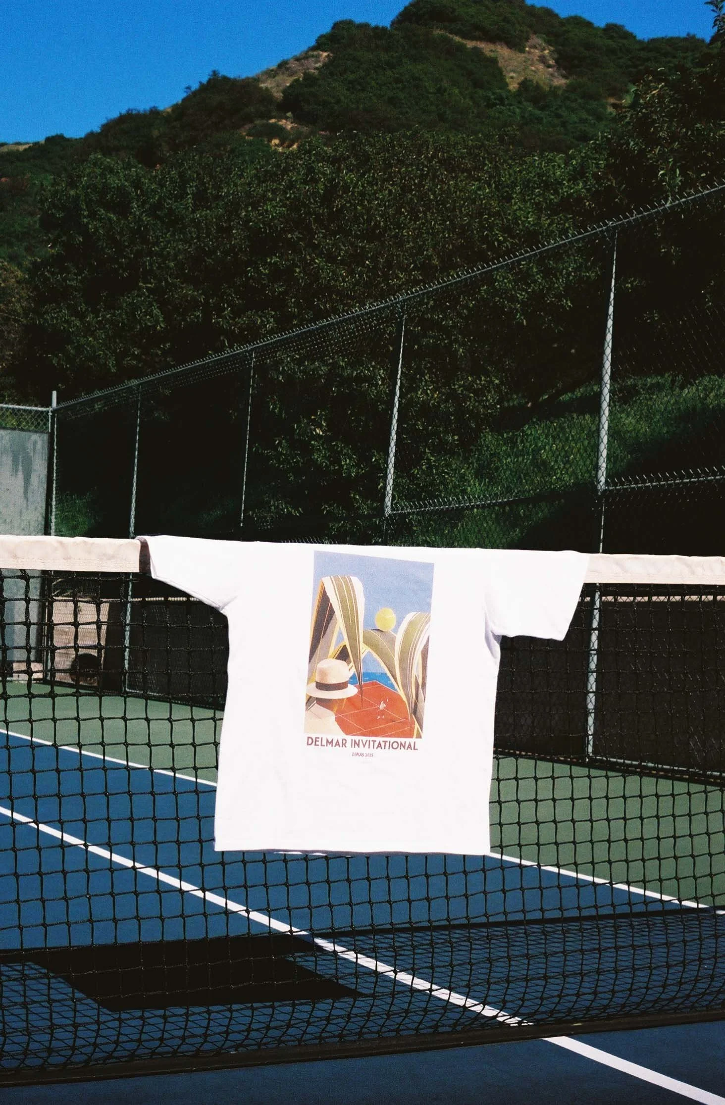 A white T-shirt with a colorful vintage-style poster design, hanging on a tennis net at an outdoor tennis court with greenery and a hill in the background.