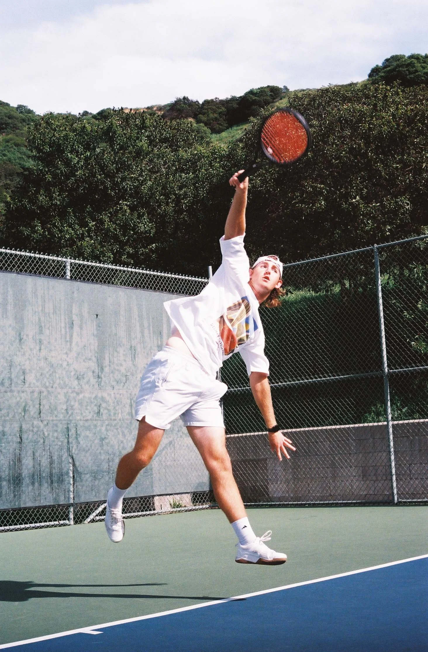 A young man with long hair playing tennis on an outdoor court, reaching up to hit a ball with a tennis racket, wearing a white t-shirt, white shorts, and white sneakers.
