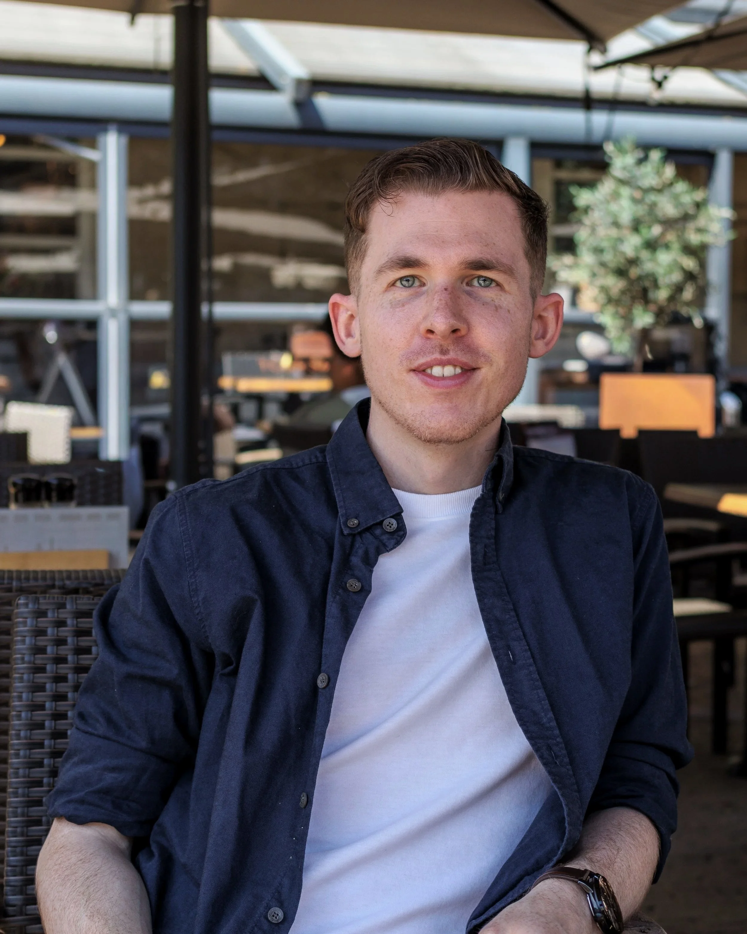 Een jonge man met korte kastanjebruine haar en blauwe ogen, zittend op een terras met rieten stoelen, onder een overkapping, lachend naar de camera.