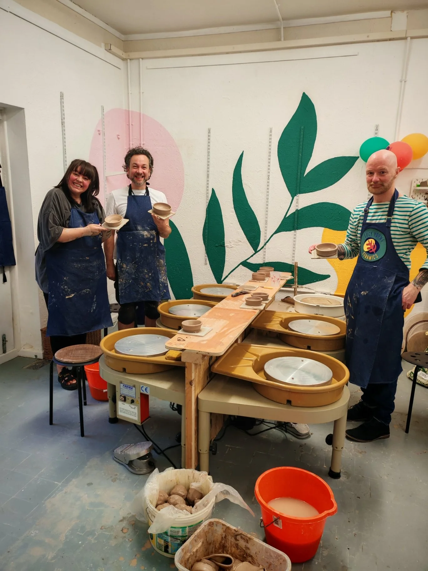 Three people in aprons and pottery gloves stand around a pottery wheel with clay, smiling and holding partially shaped bowls, in a ceramics studio decorated with large leafy wall art and colorful balloons.