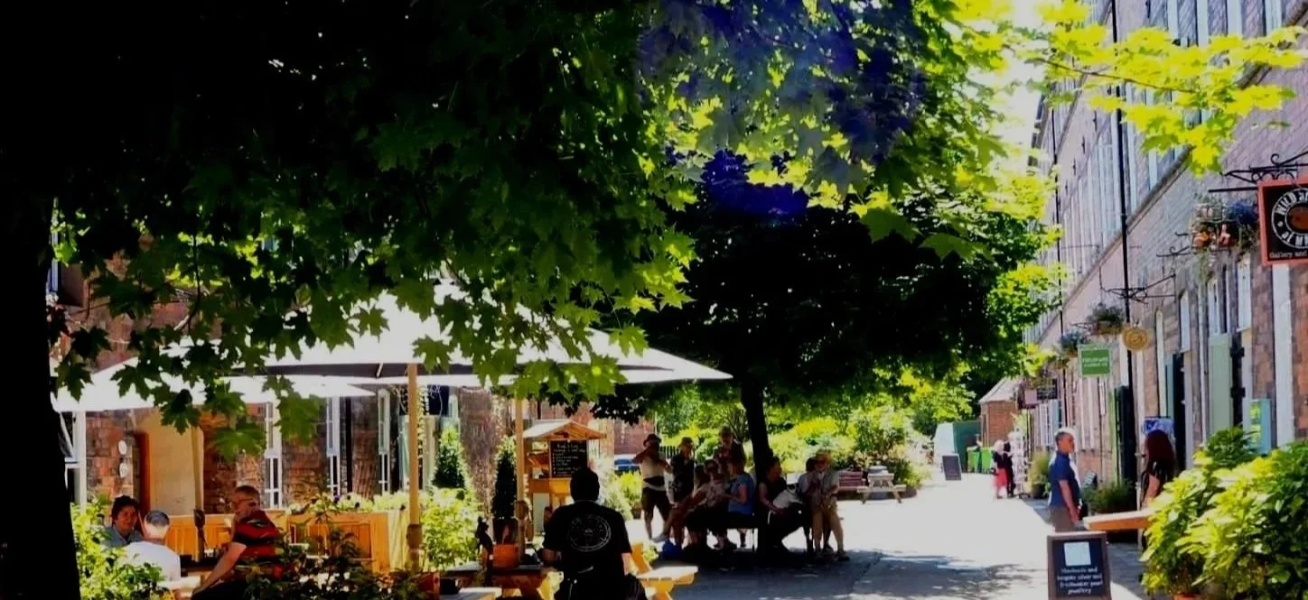 People sitting and standing at an outdoor cafe under green trees on a sunny day, with brick buildings along the street.
