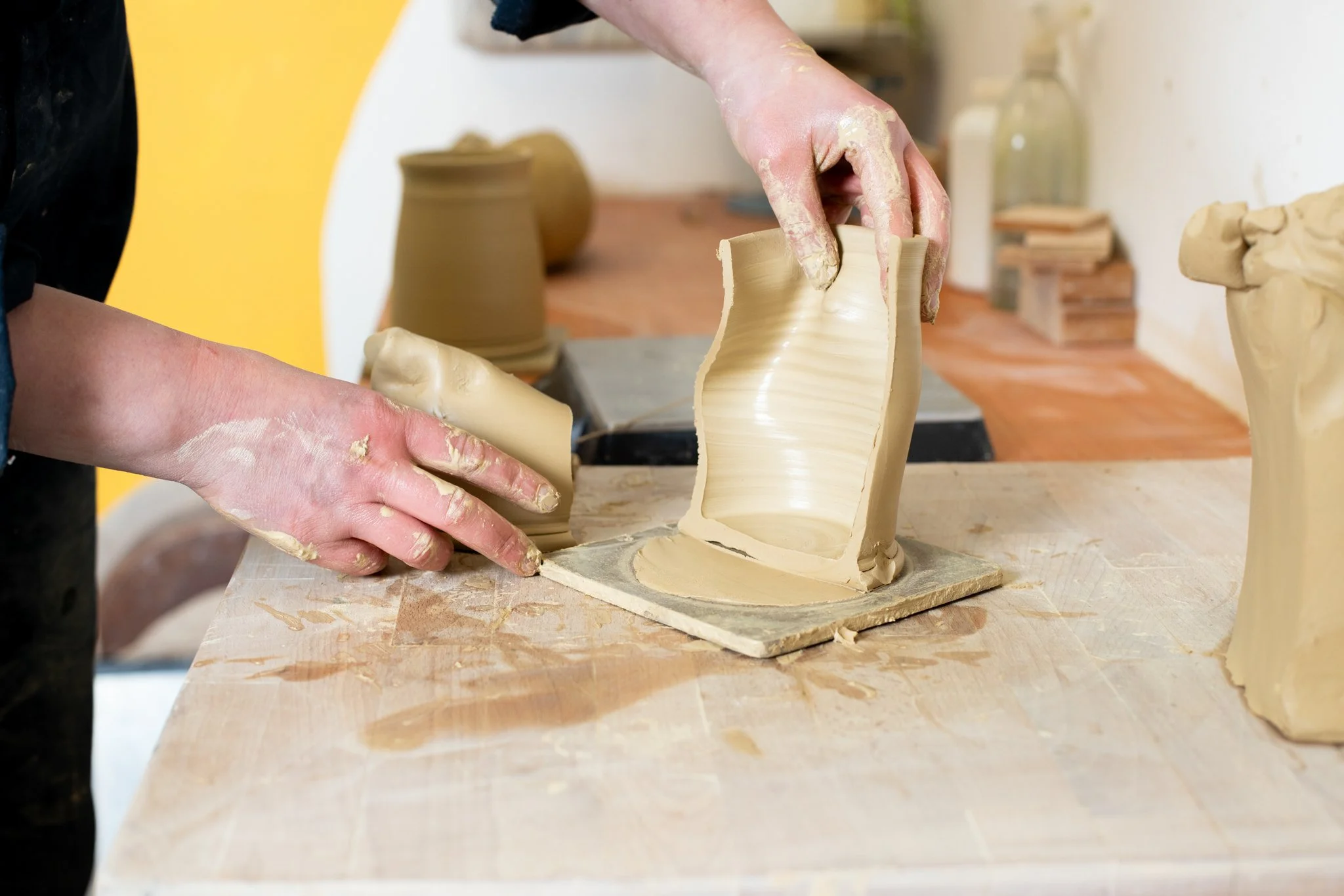 Person shaping a clay sculpture with their hands on a wooden table in a pottery studio.