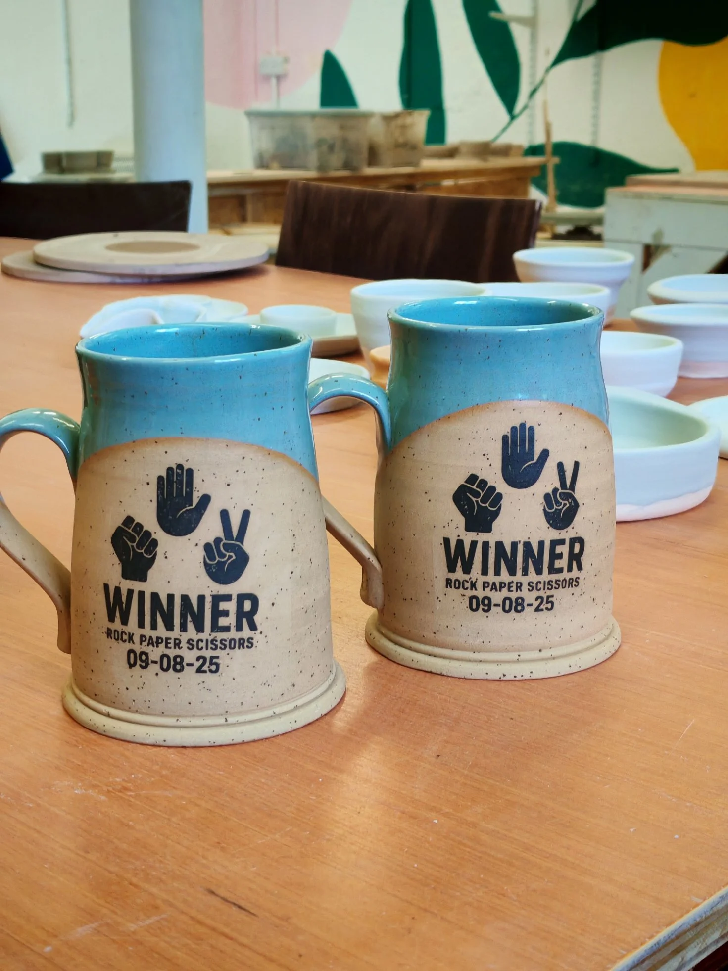 Two ceramic mugs with blue and beige glaze, featuring black text and graphics that read "WINNER ROCK PAPER SCISSORS 09-08-25" and depict hand signs for rock, paper, and scissors, sitting on a wooden table.