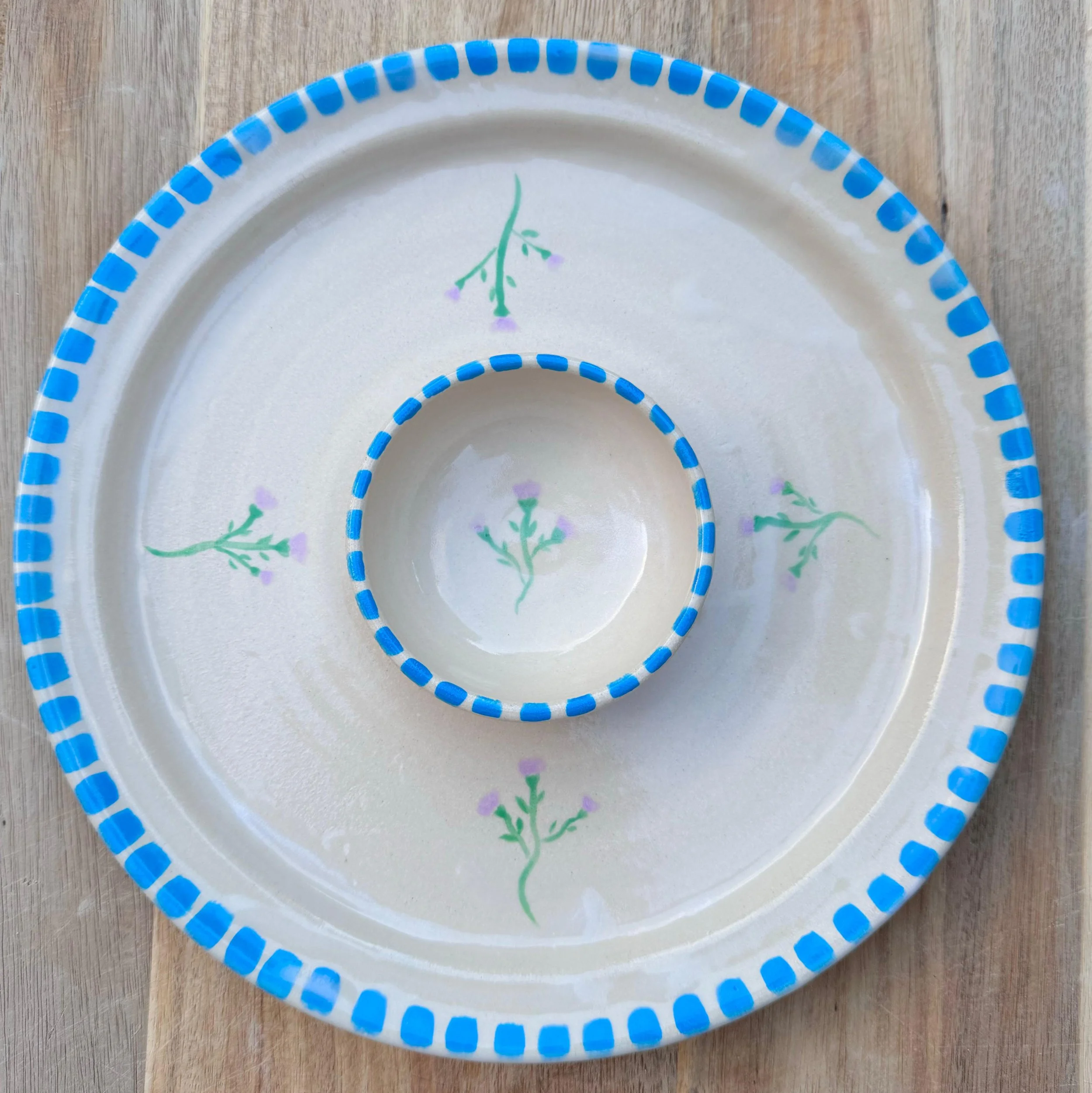 A decorative ceramic plate with a smaller bowl inside, both with blue checkered rims and hand-painted green and purple floral designs.