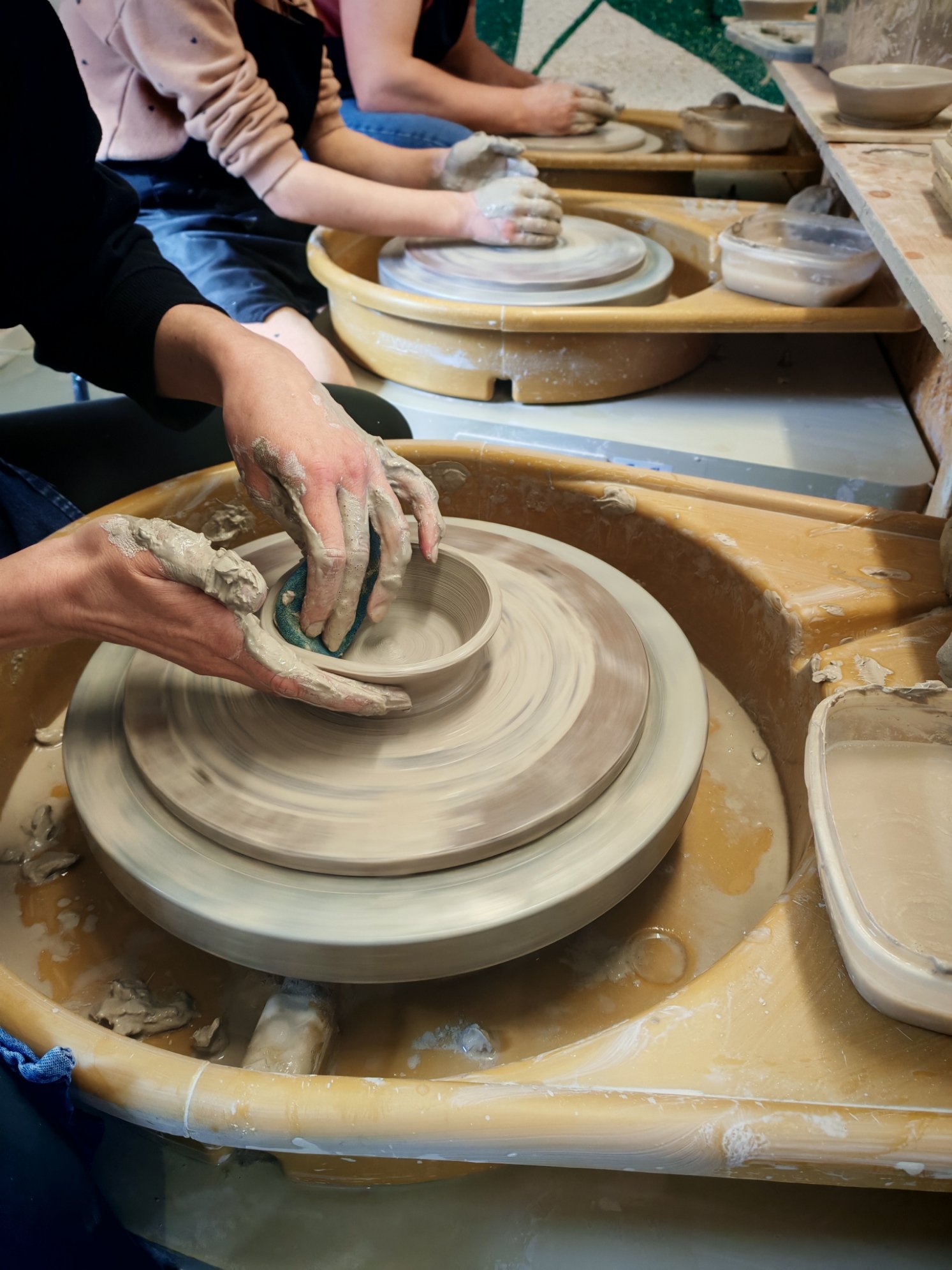 People making pottery on pottery wheels in a ceramics class, hands covered in clay and shaping bowls.
