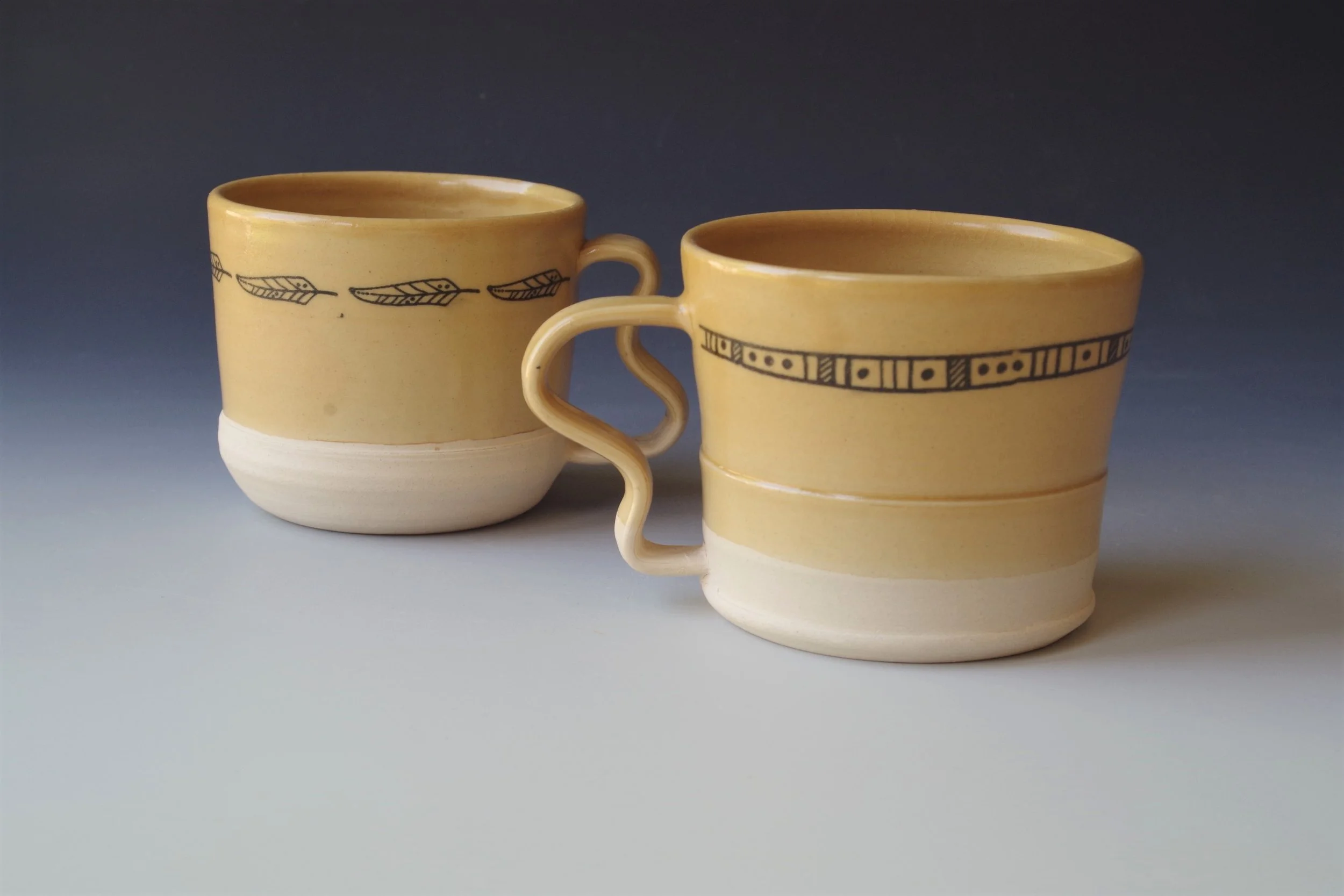 Two yellow ceramic coffee mugs with black geometric and feather patterns, one with a curved handle, placed on a light surface with a dark gradient background.