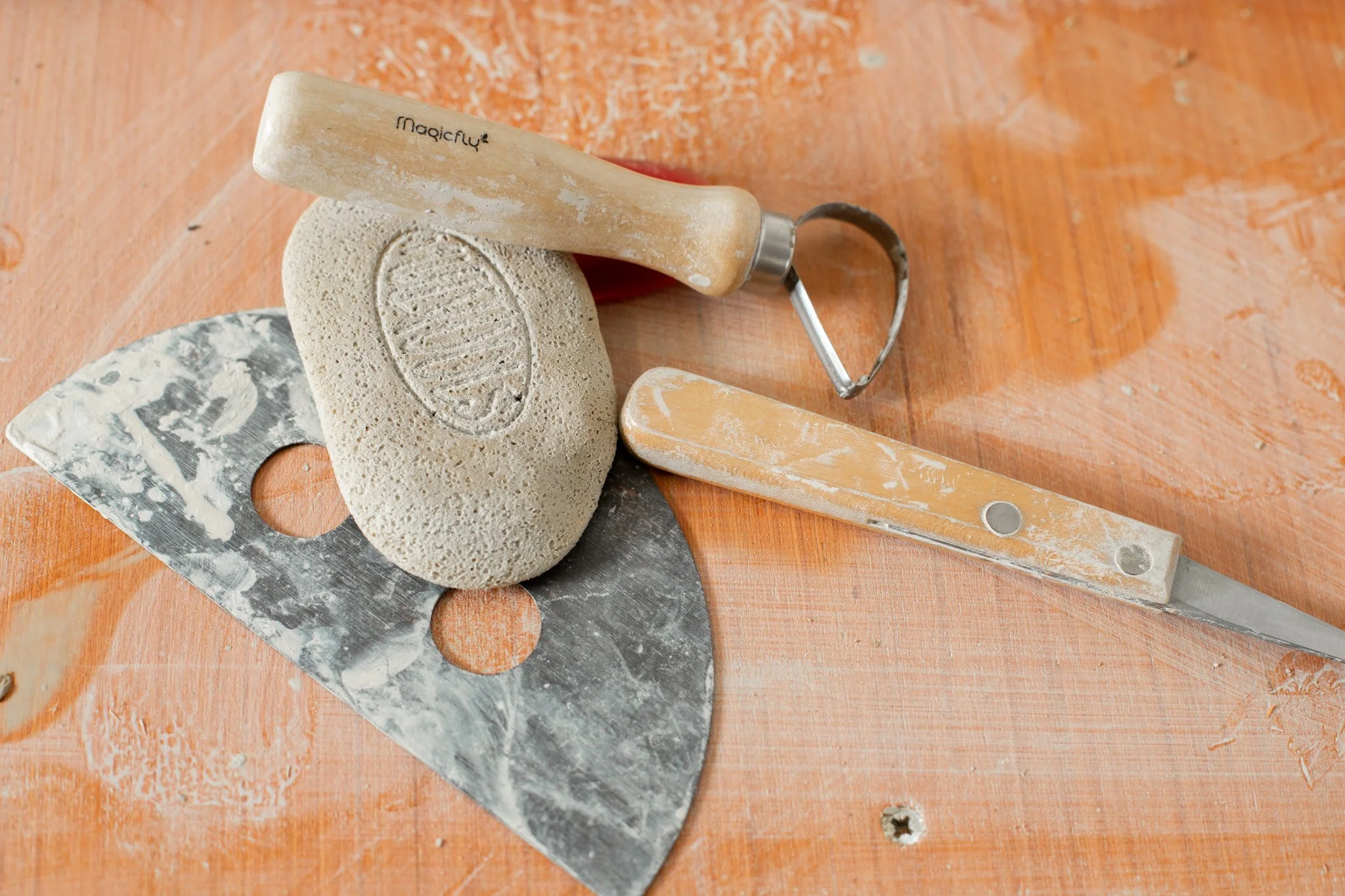 A brick of surfboard wax, a metal scraper, a sponge, and a plastic putty knife on a wooden surface, with some excess wax and marks from use.