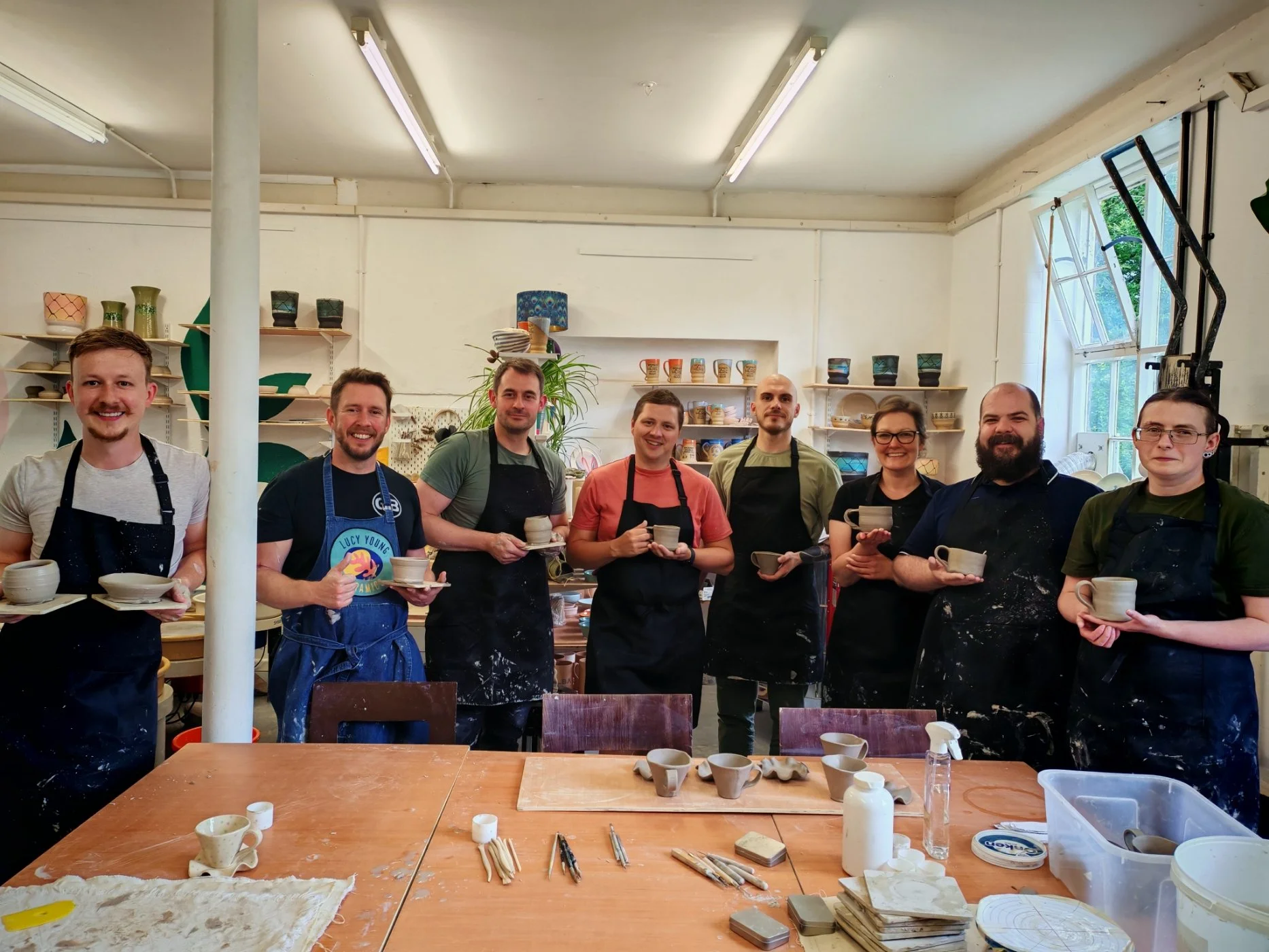 Group of nine people in a pottery workshop, holding ceramic cups they made, with pottery and tools on the table and shelves behind them.