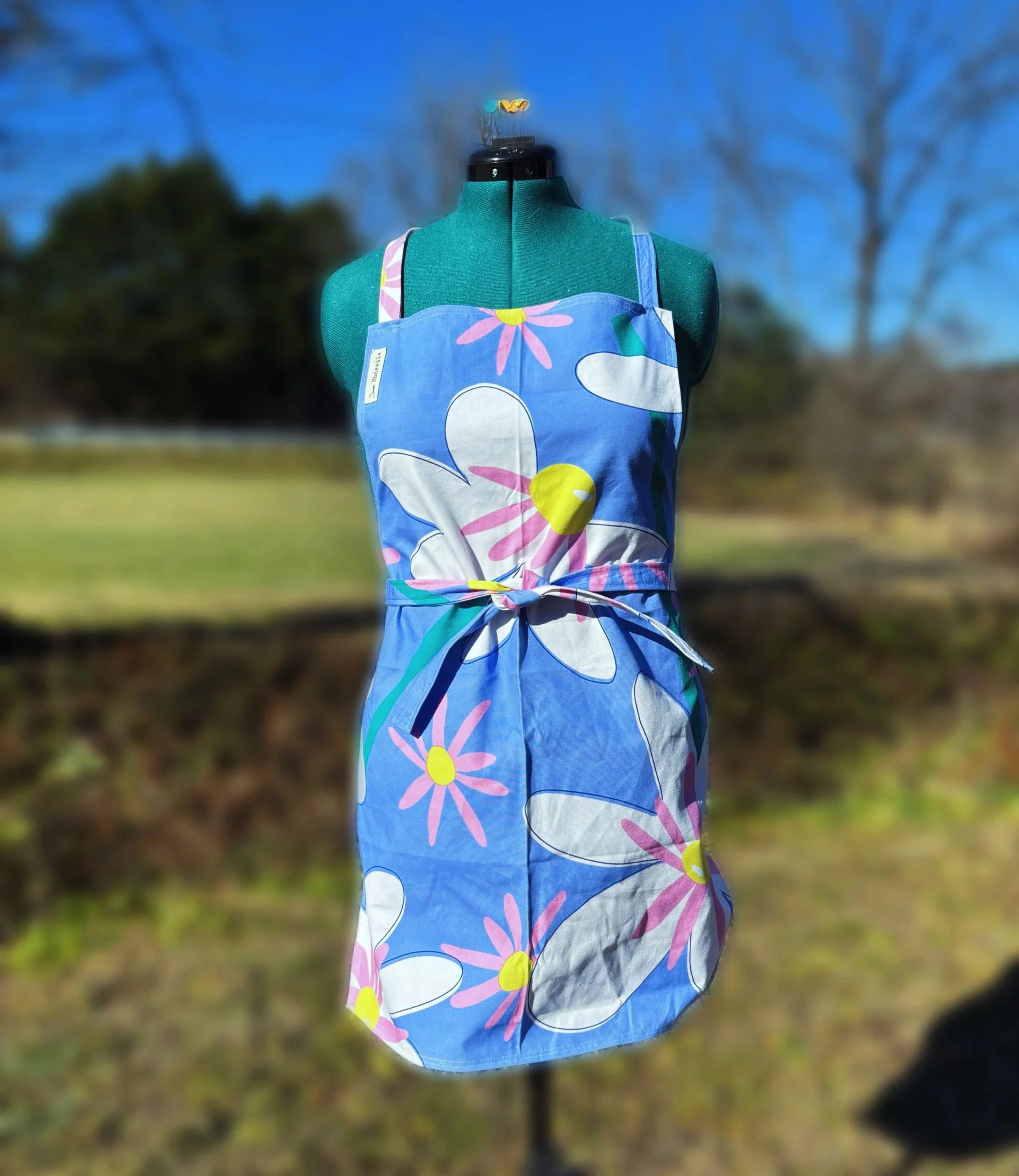 A bright sky blue apron with white and pink flowers and yellow centers. The apron is placed on a green manaquin with a blurred background of a field.