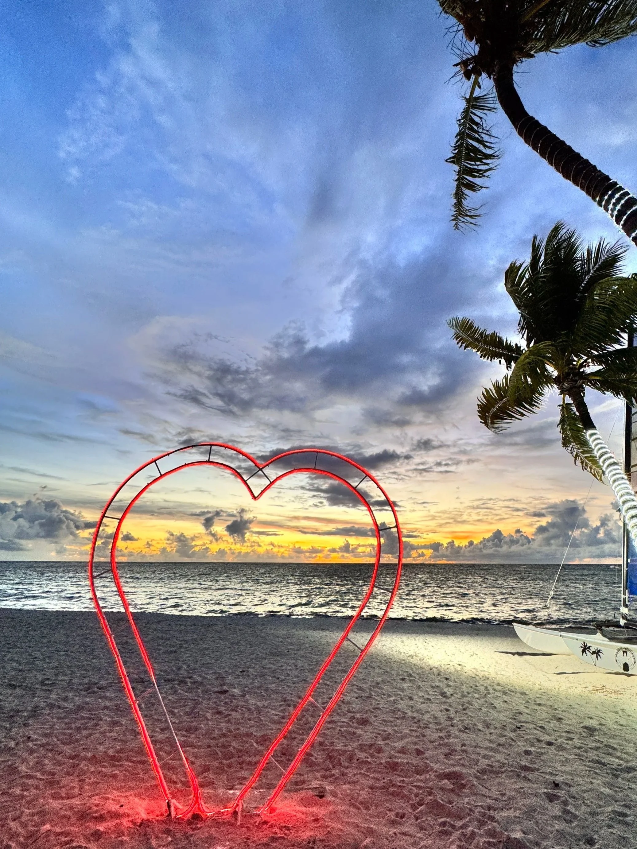 Neon red heart shape on a beach at sunset with palm trees and ocean in the background.