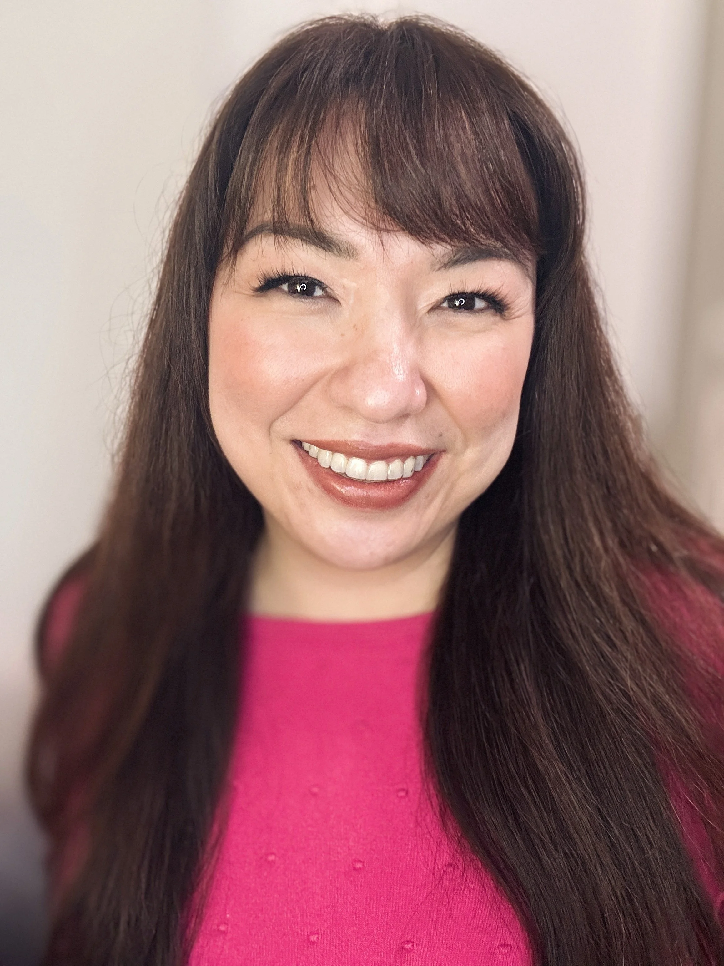 A woman with long brown hair, smiling, wearing a pink top, standing against a neutral background.