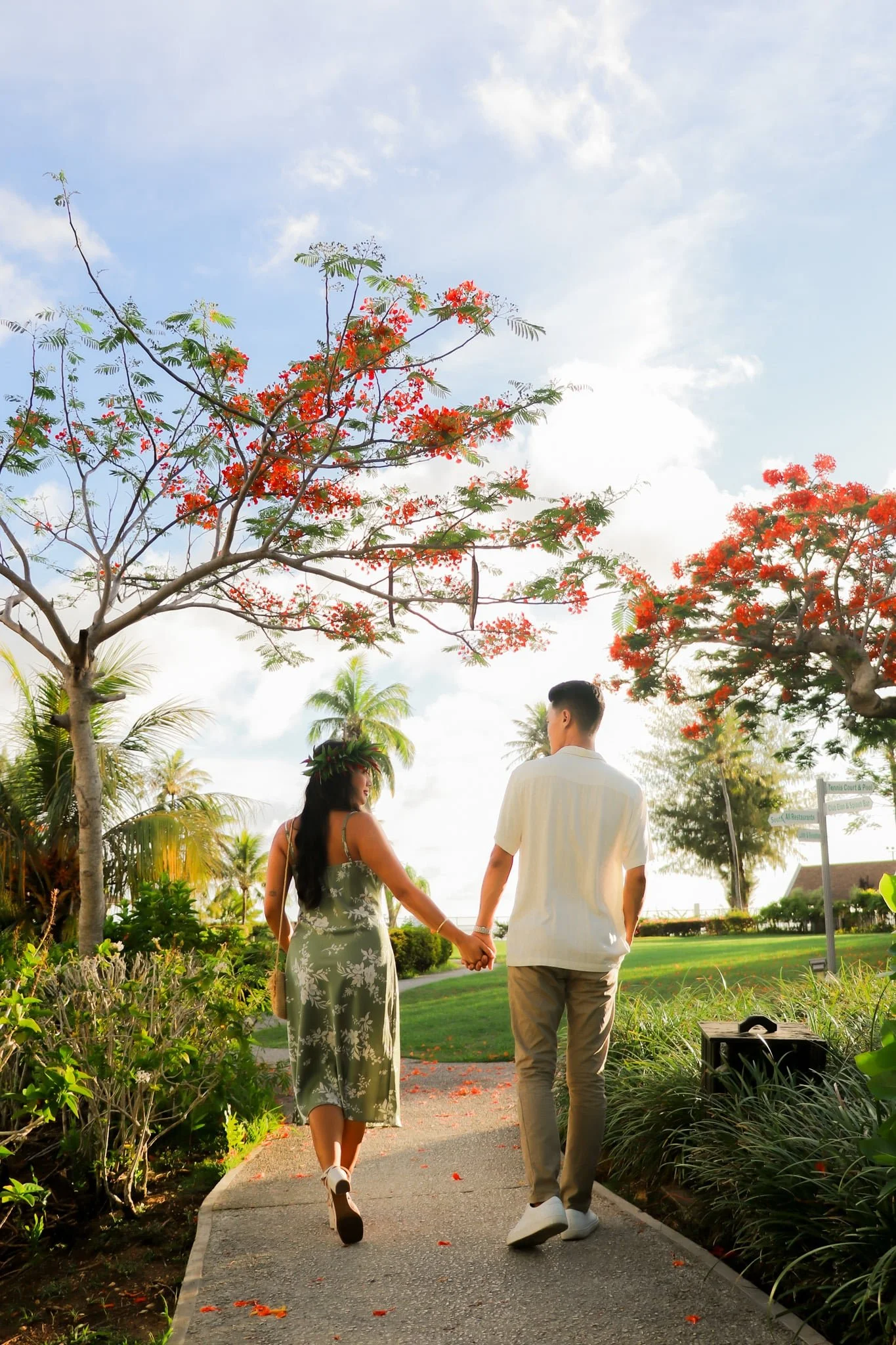 A couple walking hand in hand on a park pathway, surrounded by lush greenery and red flowering trees, under a partly cloudy sky.