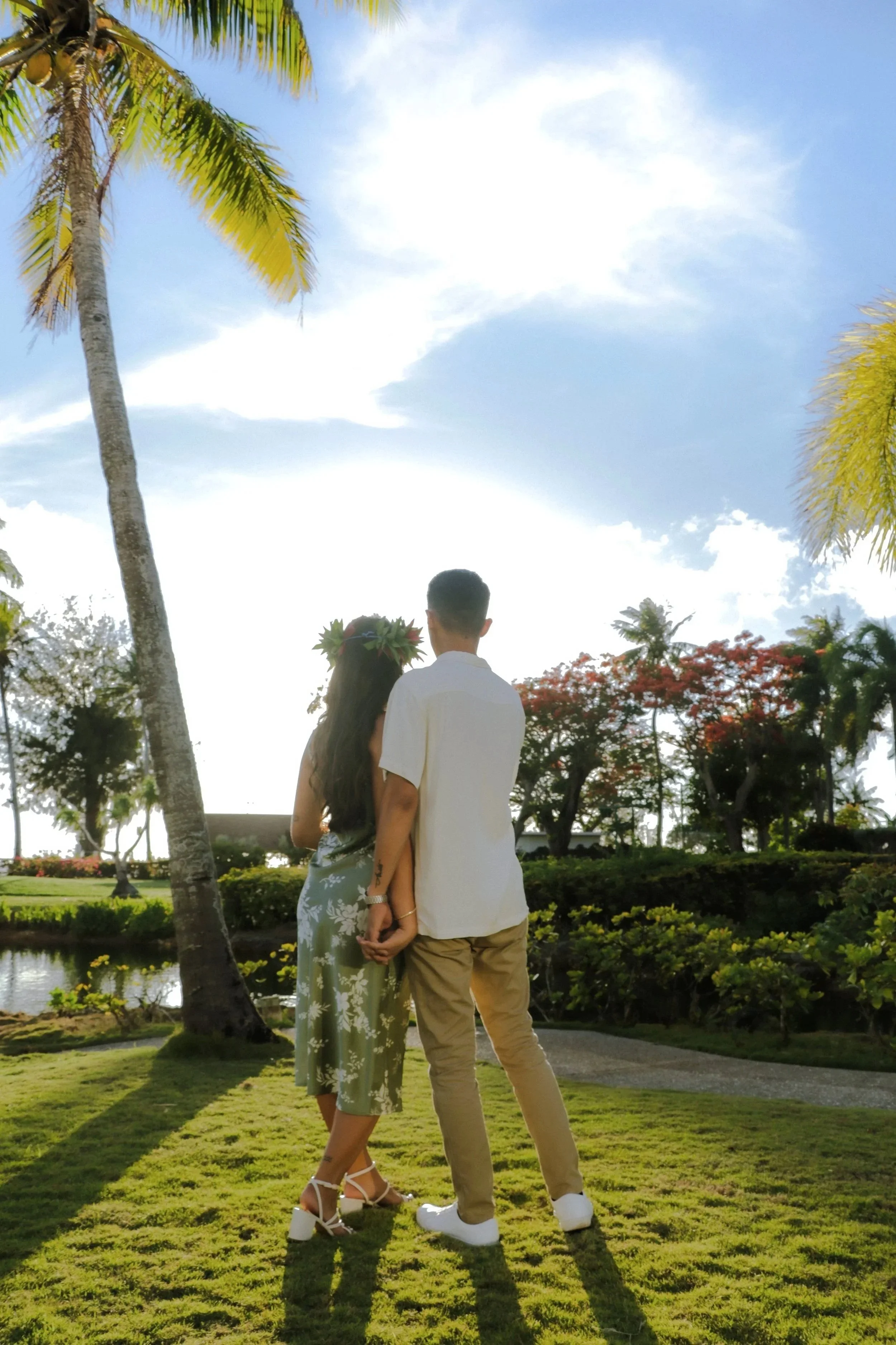 A couple standing hand in hand outdoors, facing away, in a lush garden with palm trees and vibrant plants, under a partly cloudy sky.