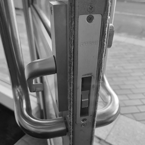 A close-up of a door lock mechanism on a glass door, showing the latch, handle, and lock plate in black and white.