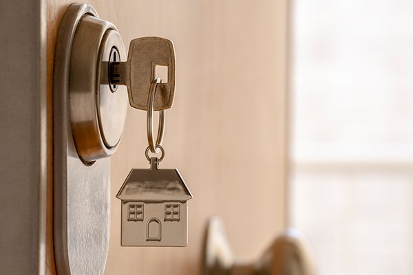 Close-up of a house-shaped keychain hanging from a house key in a door lock, with a blurred background.