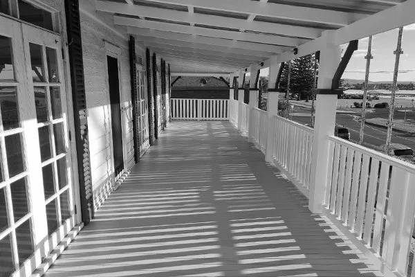 Black and white photo of a covered porch with wooden floorboards, white railing, and support beams, with houses and parking lot visible in the background.