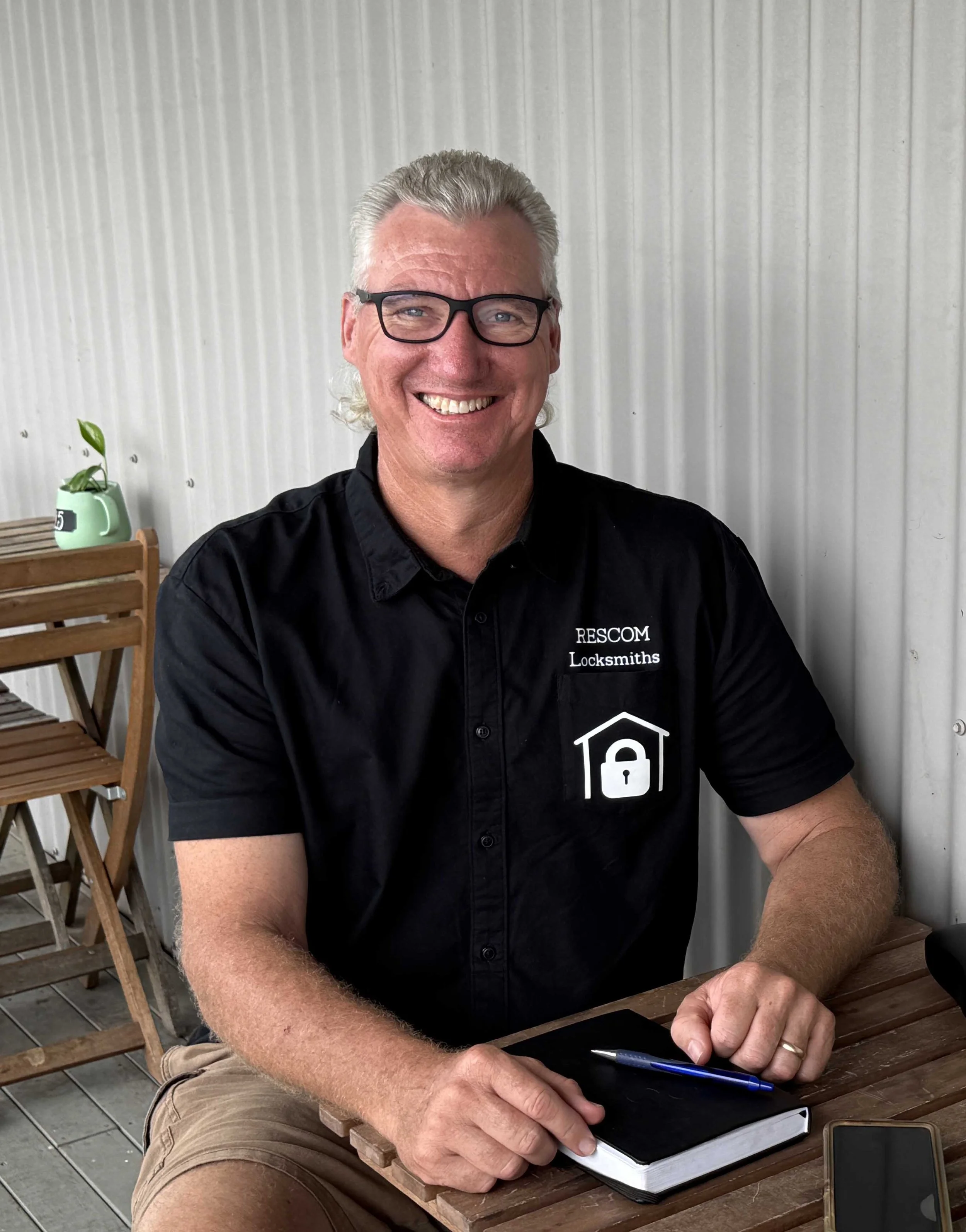 Albion Park locksmith Dan Park, sitting at a wooden table with a notebook, pen, and smartphone, planning his next job.