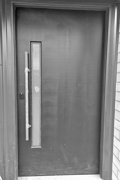 A black and white image of a wooden front door with a vertical window and a vertical handle, flanked by brick wall on the right side.