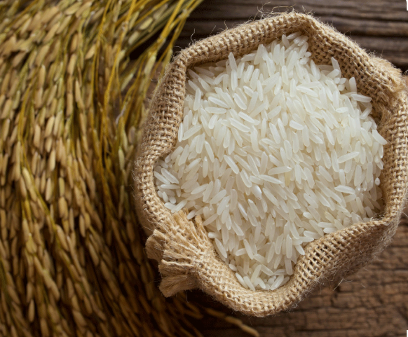 A burlap sack filled with white rice on a wooden surface, with a woven straw mat partially visible on the side. Jasmine rice, dinorado, dikit, glutinous rice, and others