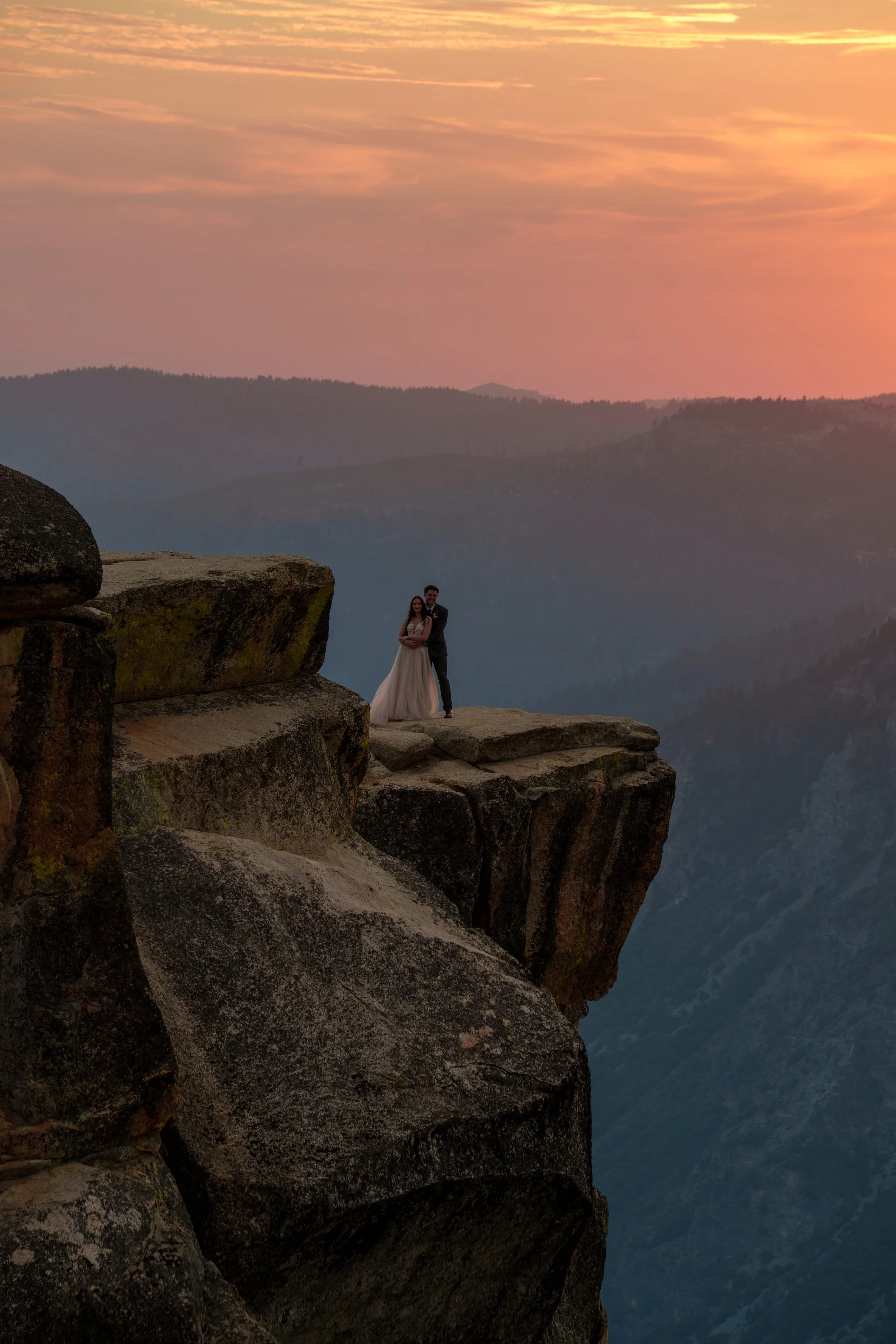 A couple dressed in wedding attire standing on a rocky ledge overlooking a mountainous landscape during sunset. The sky is painted with pink and orange hues.