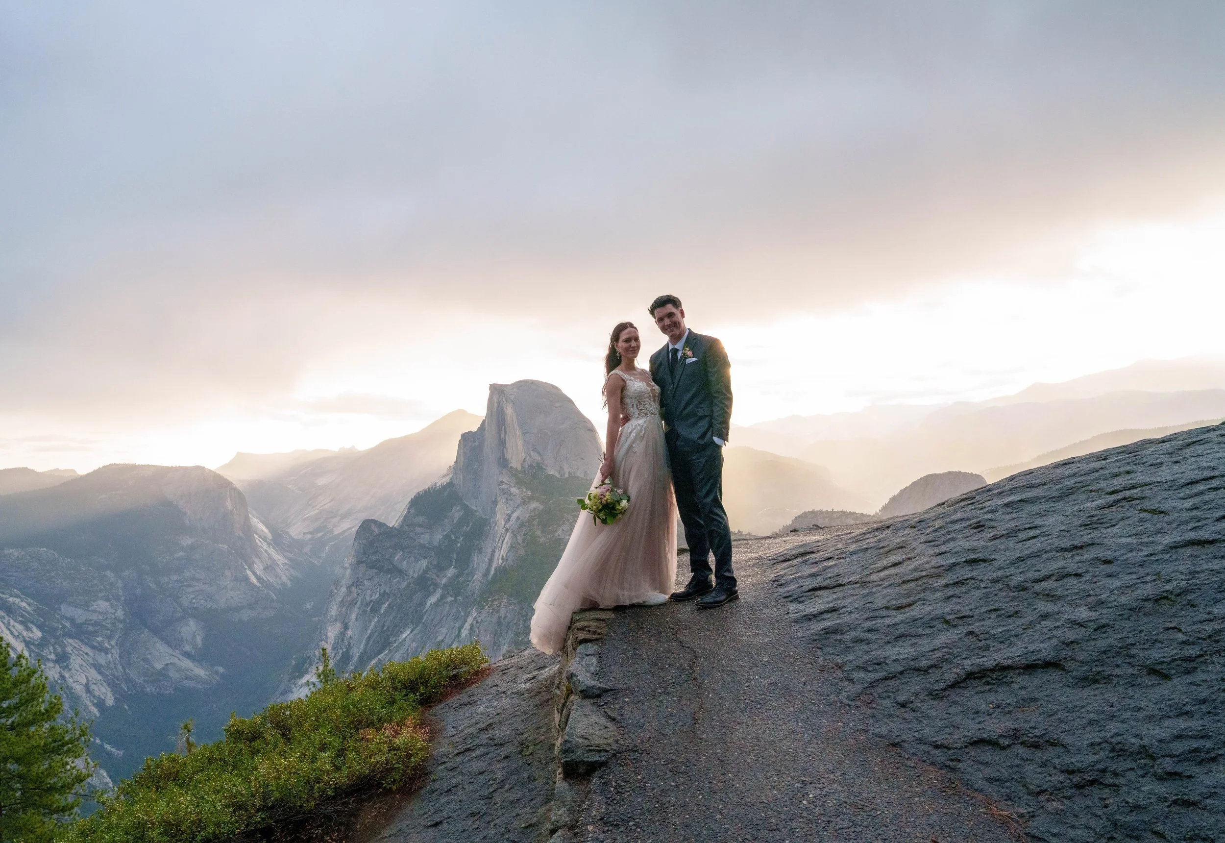 A bride and groom standing on a rocky outcrop with a mountainous landscape in the background during sunset. Yosemite Nation Park. Yosemite Elopement Photography. Yosemite Elopement Videography