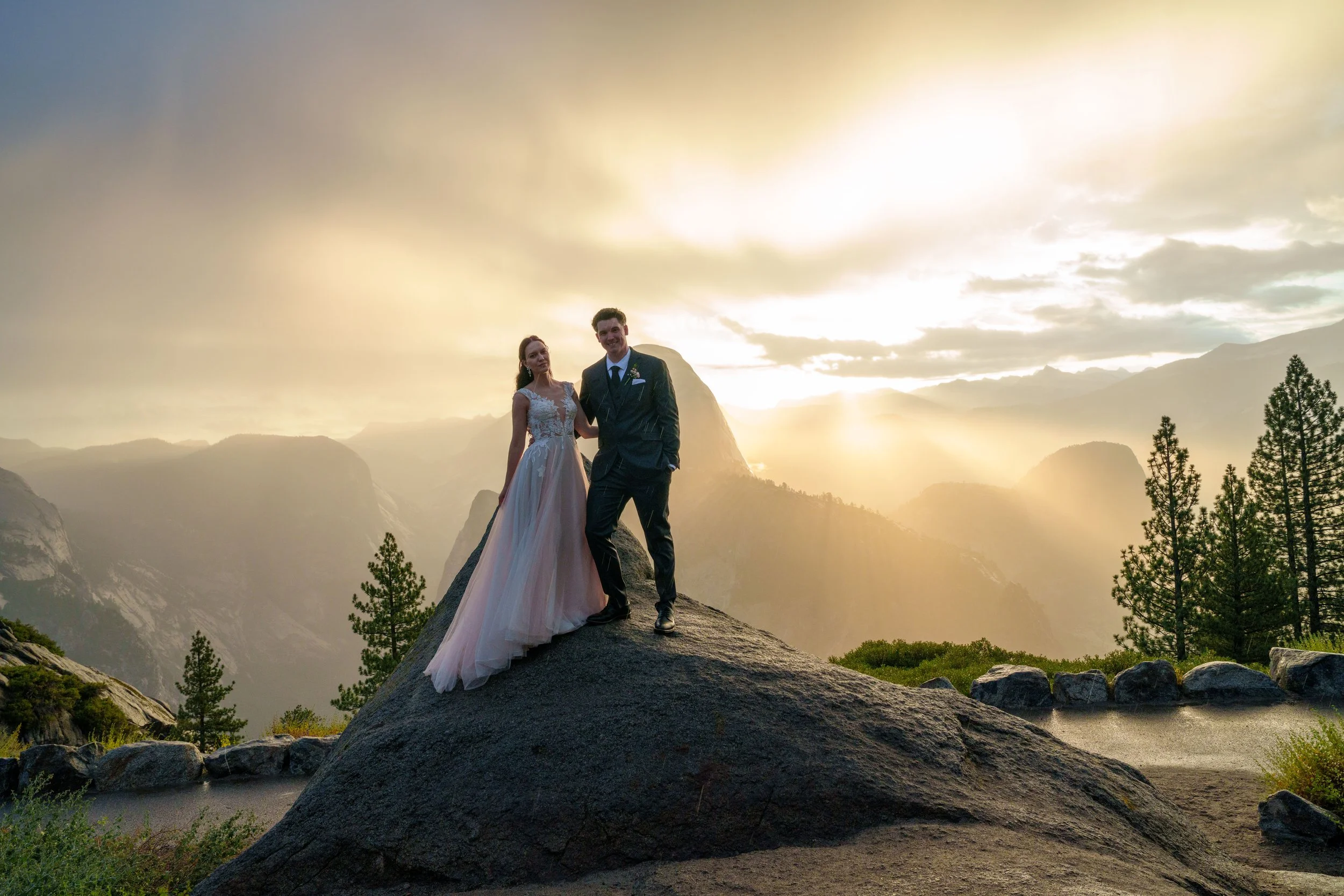 A bride and groom standing on a large rock in a mountainous landscape during sunset, with trees in the foreground and mountains in the background. Yosemite Nation Park. Yosemite Elopement Photography. Yosemite Elopement Videography