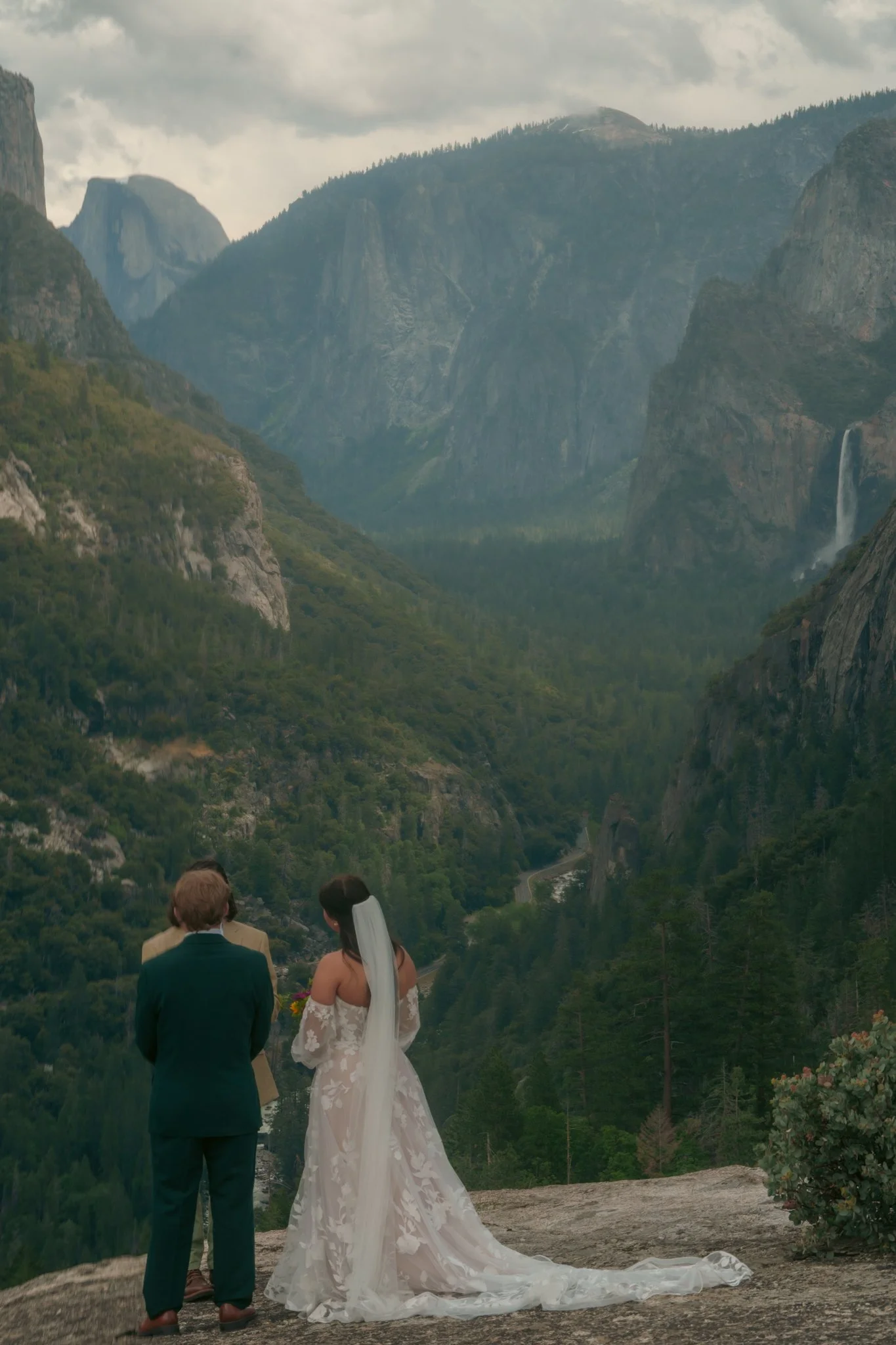 A wedding couple standing outdoors on a rocky area with a mountainous landscape background. The groom is wearing a dark suit and the bride is in a white lace wedding dress with a long train. The scenery includes large cliffs, green trees, and a water