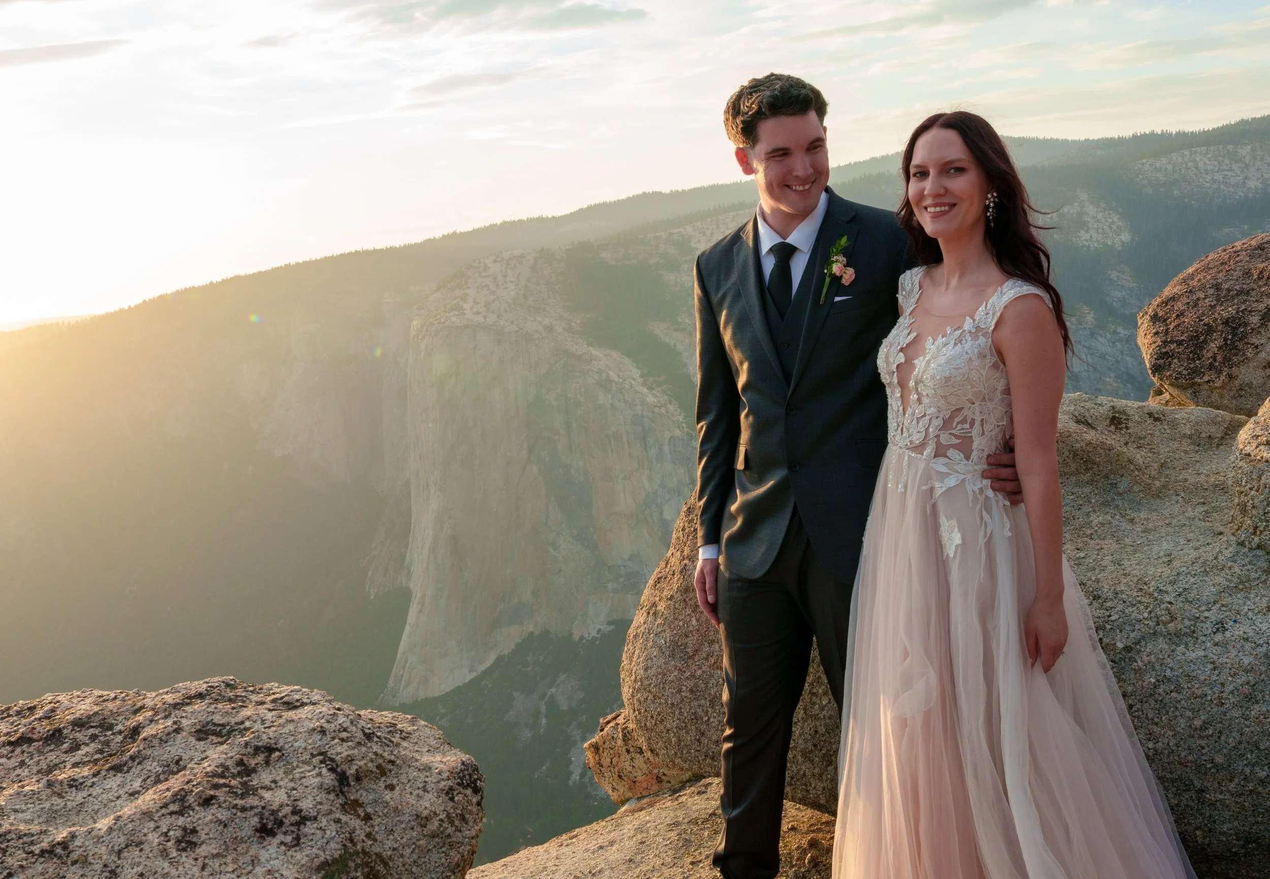 A newlywed couple standing on a rocky ledge overlooking a canyon at sunset, smiling at the camera. Yosemite Nation Park. Yosemite Elopement Photography. Yosemite Elopement Videography El Capitan, Taft Point
