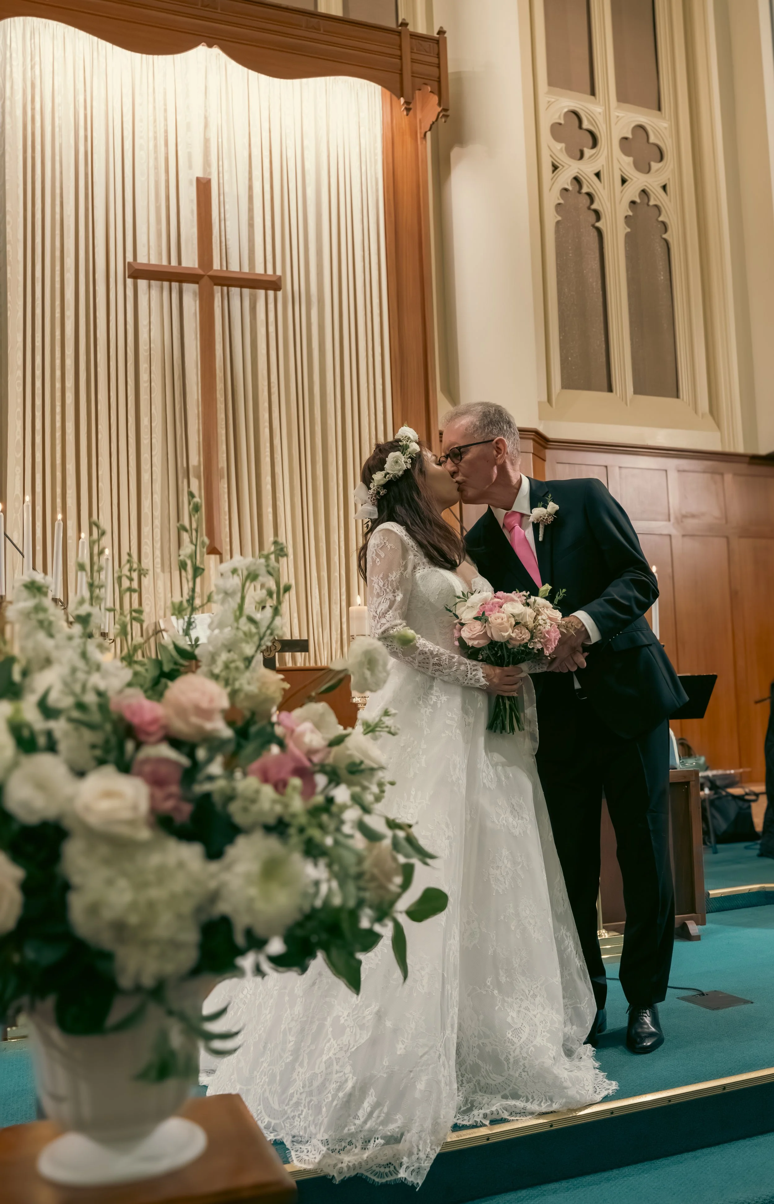 A bride and a man, possibly her father, are sharing a kiss during a wedding ceremony in a church. The bride wears a white lace wedding dress and a floral crown, and holds a bouquet of pink and white flowers. The groom or officiant is dressed in a bla