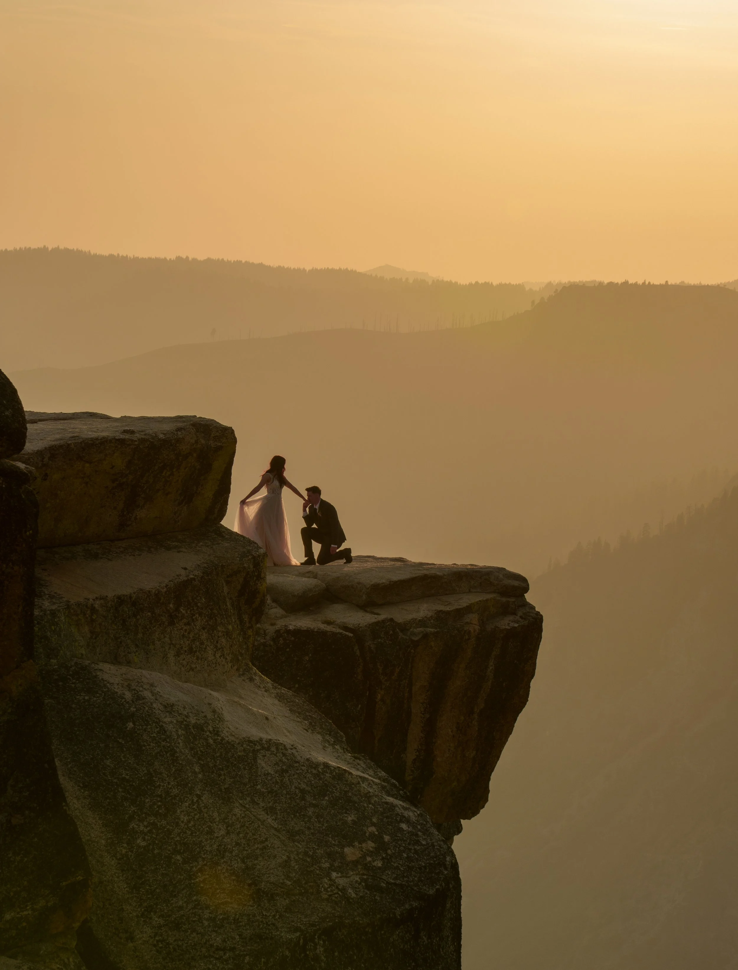 A wedding couple standing on a rocky ledge at sunset, with a woman in a wedding dress and a man in a suit, overlooking a misty valley and rolling hills. Yosemite Nation Park. Yosemite Elopement Photography. Yosemite Elopement Videography Taft Point