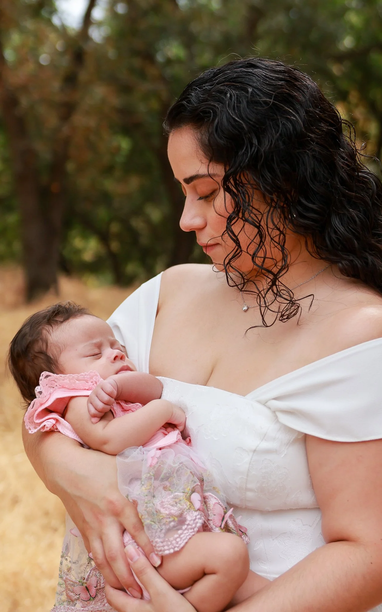 A woman holding a sleeping baby in an outdoor setting with trees in the background.