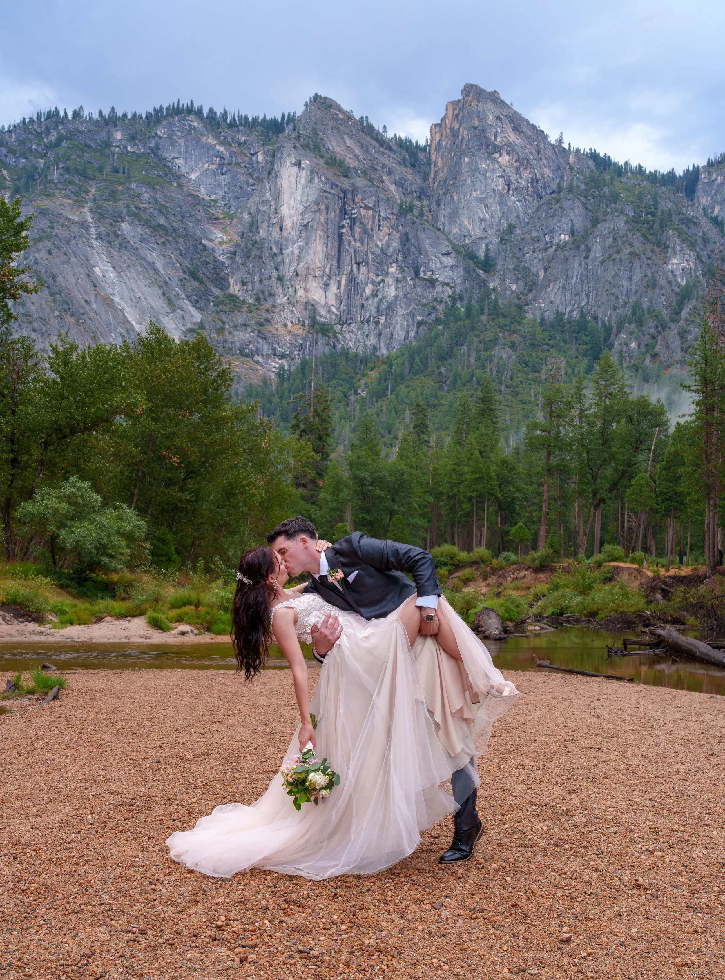 A bride and groom kissing in a scenic outdoor setting with tall trees, a river, and mountains in the background. Yosemite Nation Park. Yosemite Elopement Photography. Yosemite Elopement Videography Cathedral Beach