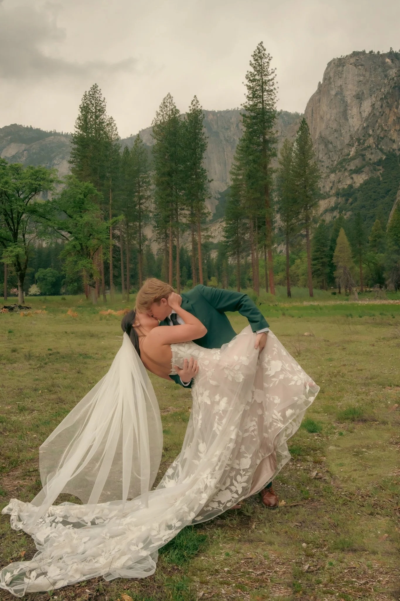 A couple in wedding attire kissing outdoors in a grassy field with green trees and mountains in the background.
