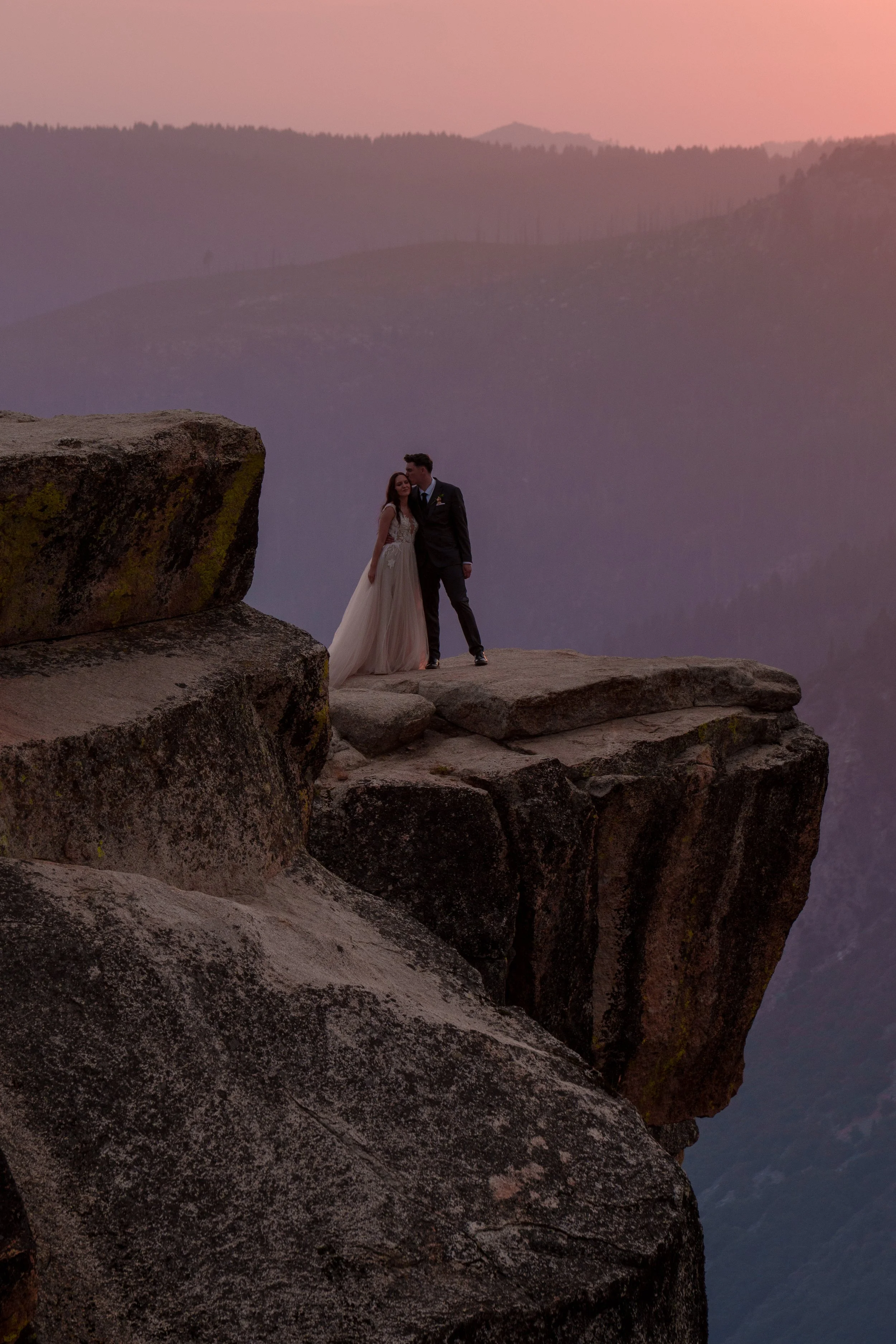 A bride and groom standing close together on a rocky cliff with a mountain landscape and colorful sky in the background. Yosemite Nation Park. Yosemite Elopement Photography. Yosemite Elopement Videography Taft Point