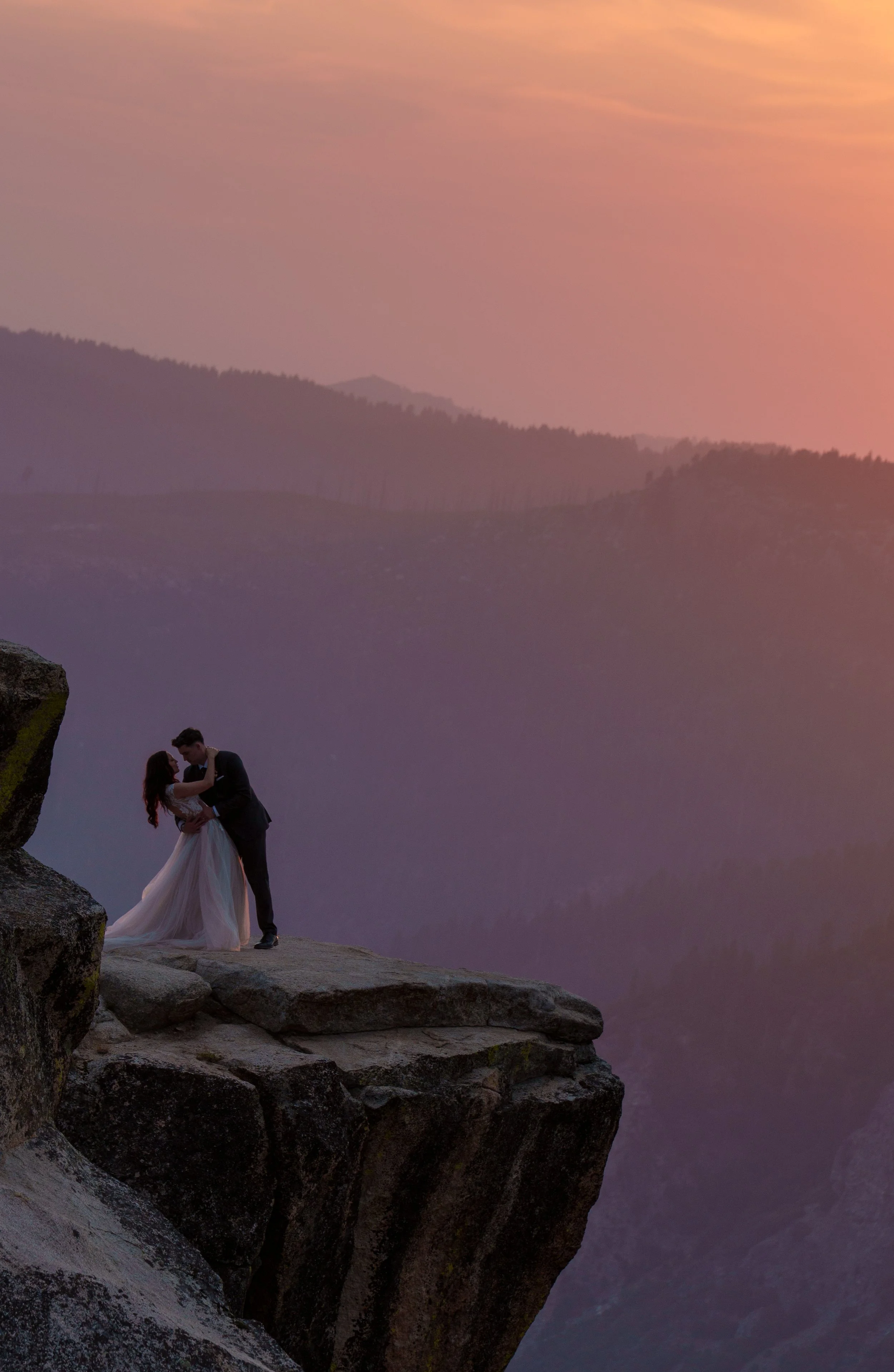 A couple dressed in wedding attire stands on a rocky cliff at sunset, with a purple and pink sky and mountain silhouettes in the background. Yosemite Nation Park. Yosemite Elopement Photography. Yosemite Elopement Videography Taft Point