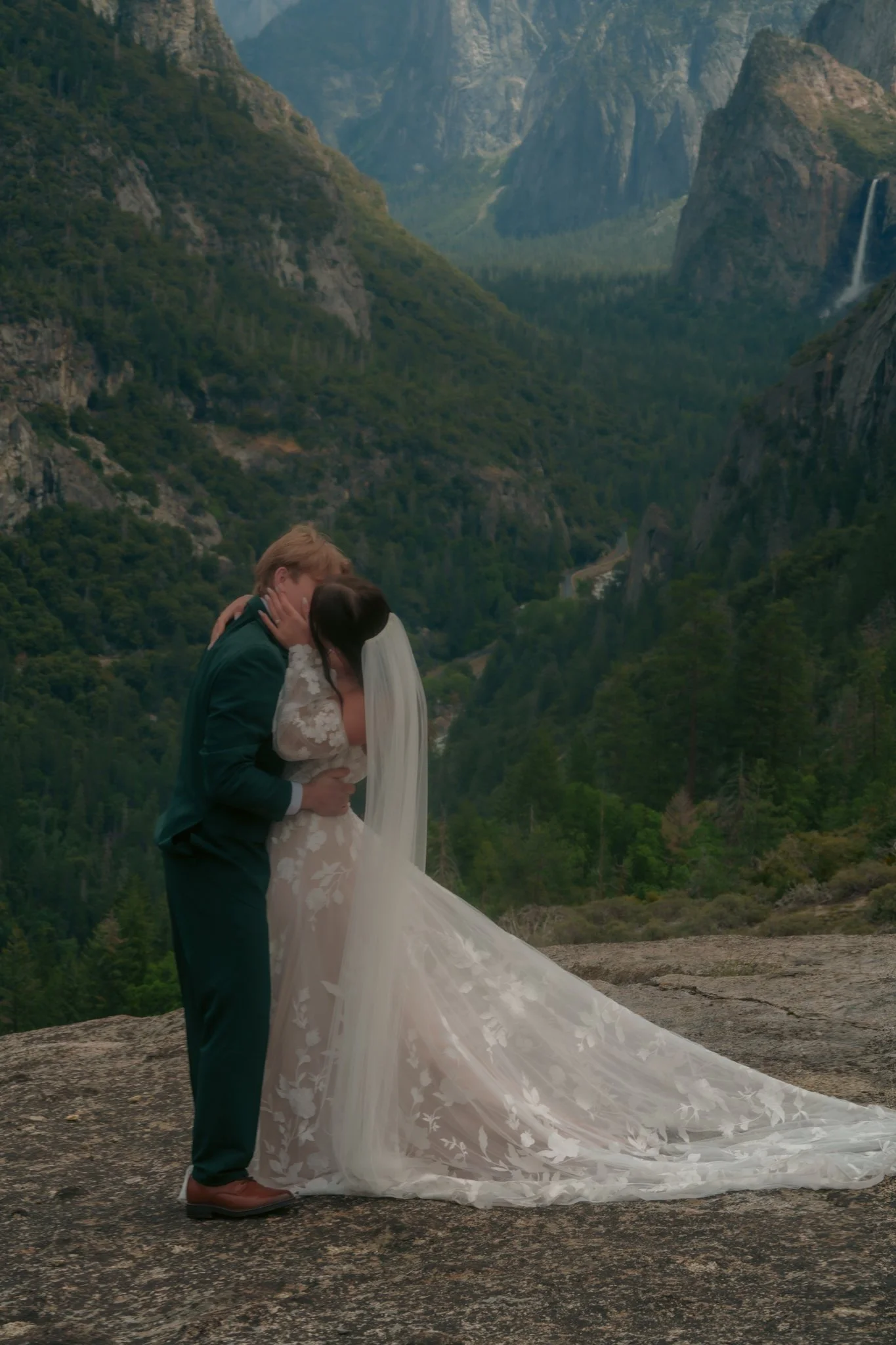 A bride and groom kissing in a mountainous landscape during their wedding, with lush green hills, cliffs, and waterfalls in the background.