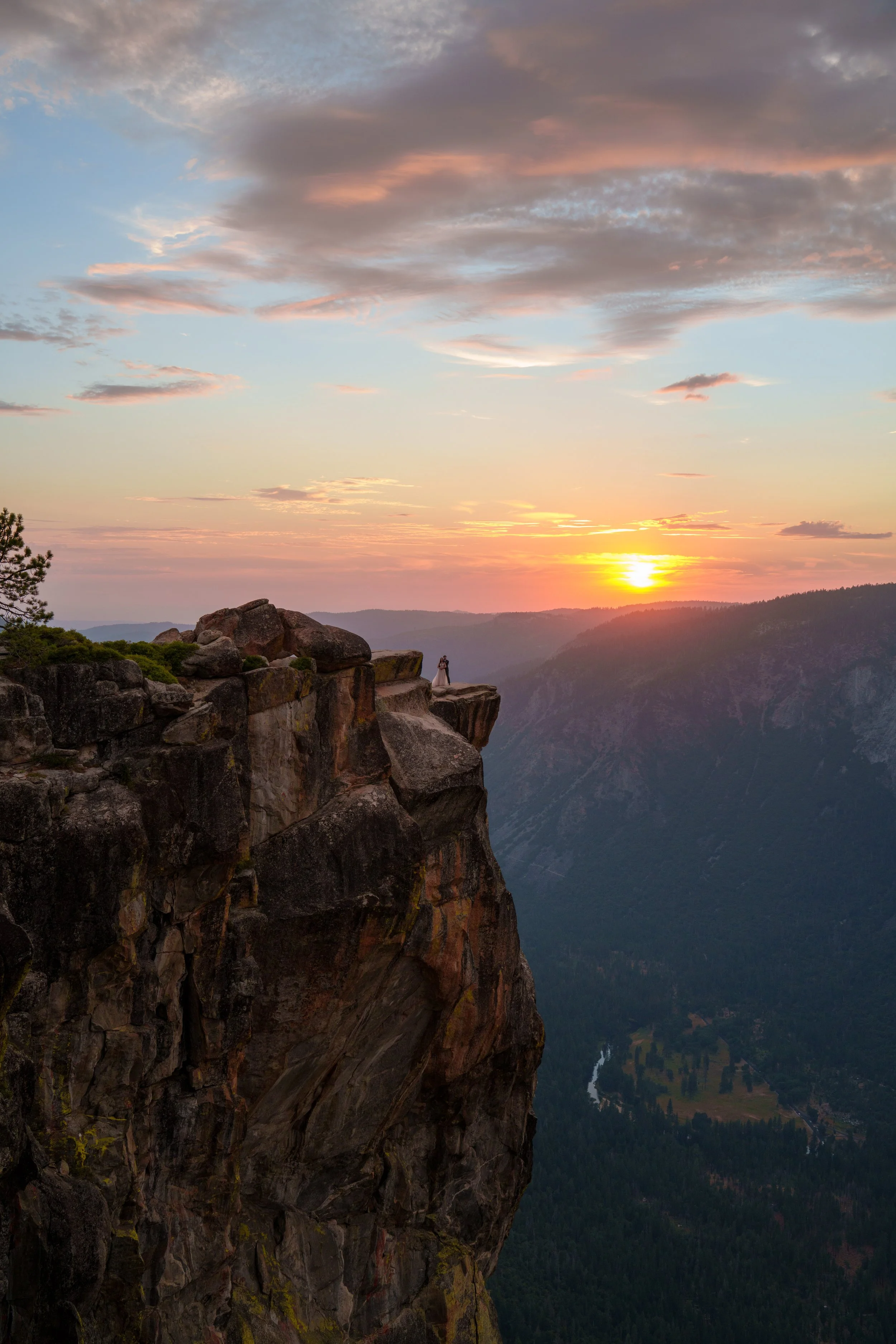 Sunset over a canyon with rocky cliffs in the foreground and a small couple standing on the edge, with colorful sky and clouds in the background. Yosemite Nation Park. Yosemite Elopement Photography. Yosemite Elopement Videography Taft Point